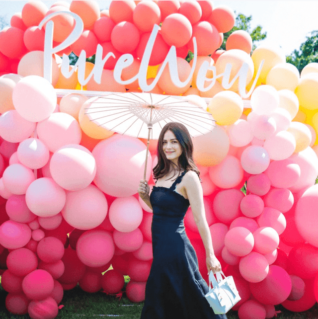 A woman holding an umbrella in front of a wall of pink balloons with the text PureWow.