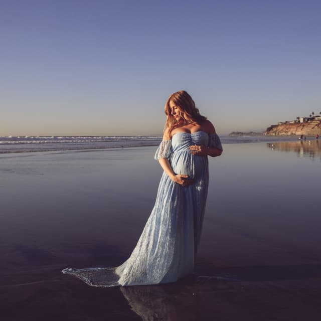 A pregnant woman standing on the beach at sunset, wearing a long blue dress.