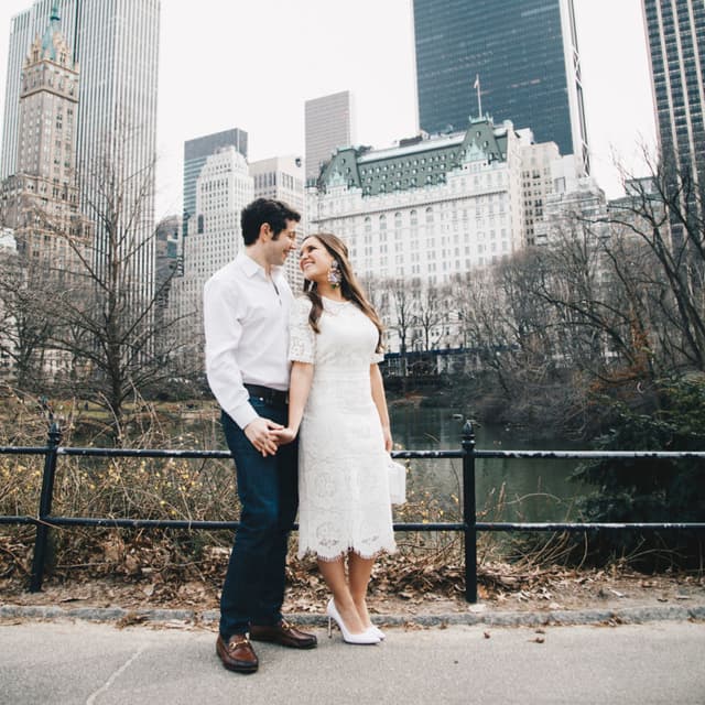 A couple holding hands and looking into each others eyes in Central Park with skyline and trees in the background.
