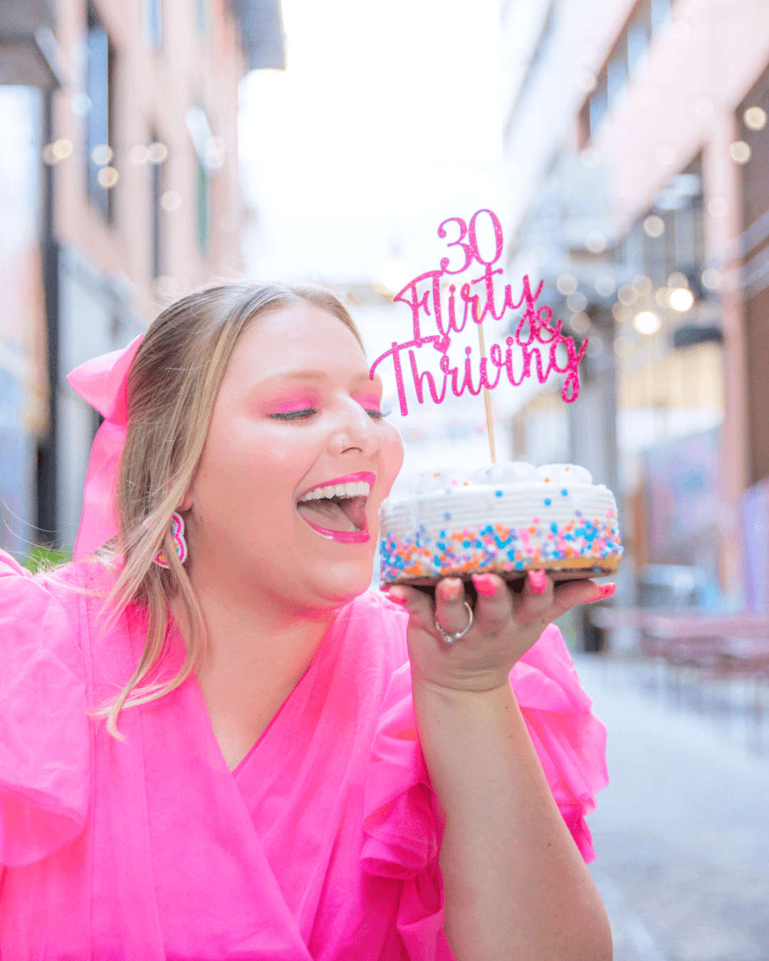 Woman in pink holding a '30 Flirty and Thriving' birthday cake in a city alley