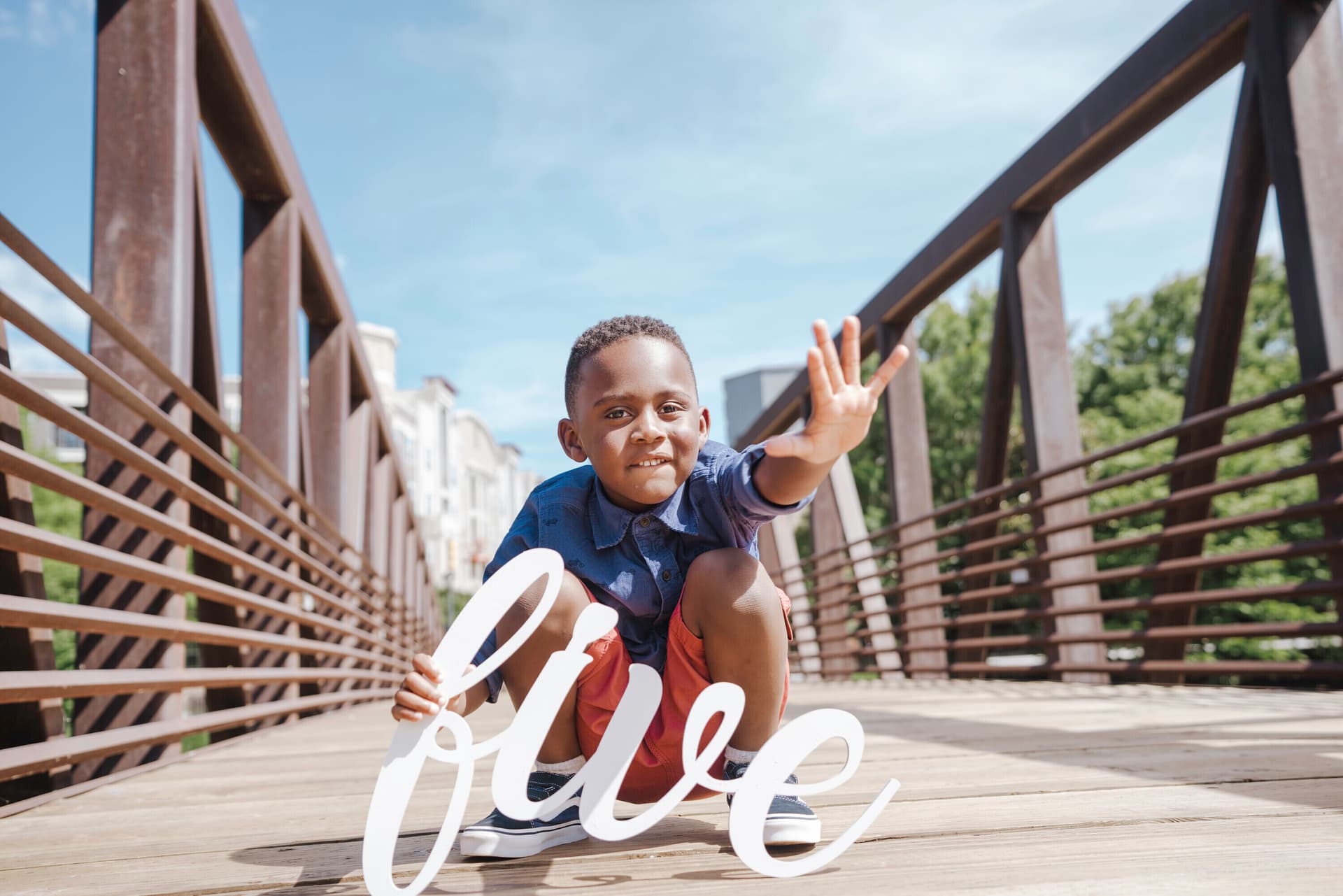 Young boy posing on a wooden bridge with a 'five' script sign celebrating his fifth birthday