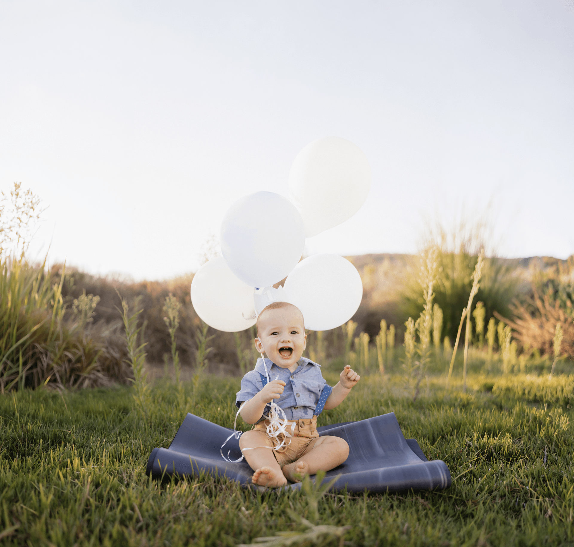 Happy baby sitting on a blanket in a golden field holding white balloons and laughing joyfully