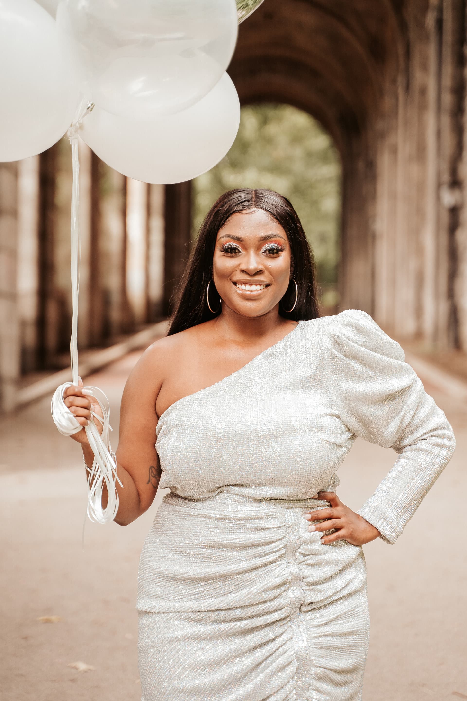 Woman in a silver sequin dress holding white balloons under a stone archway