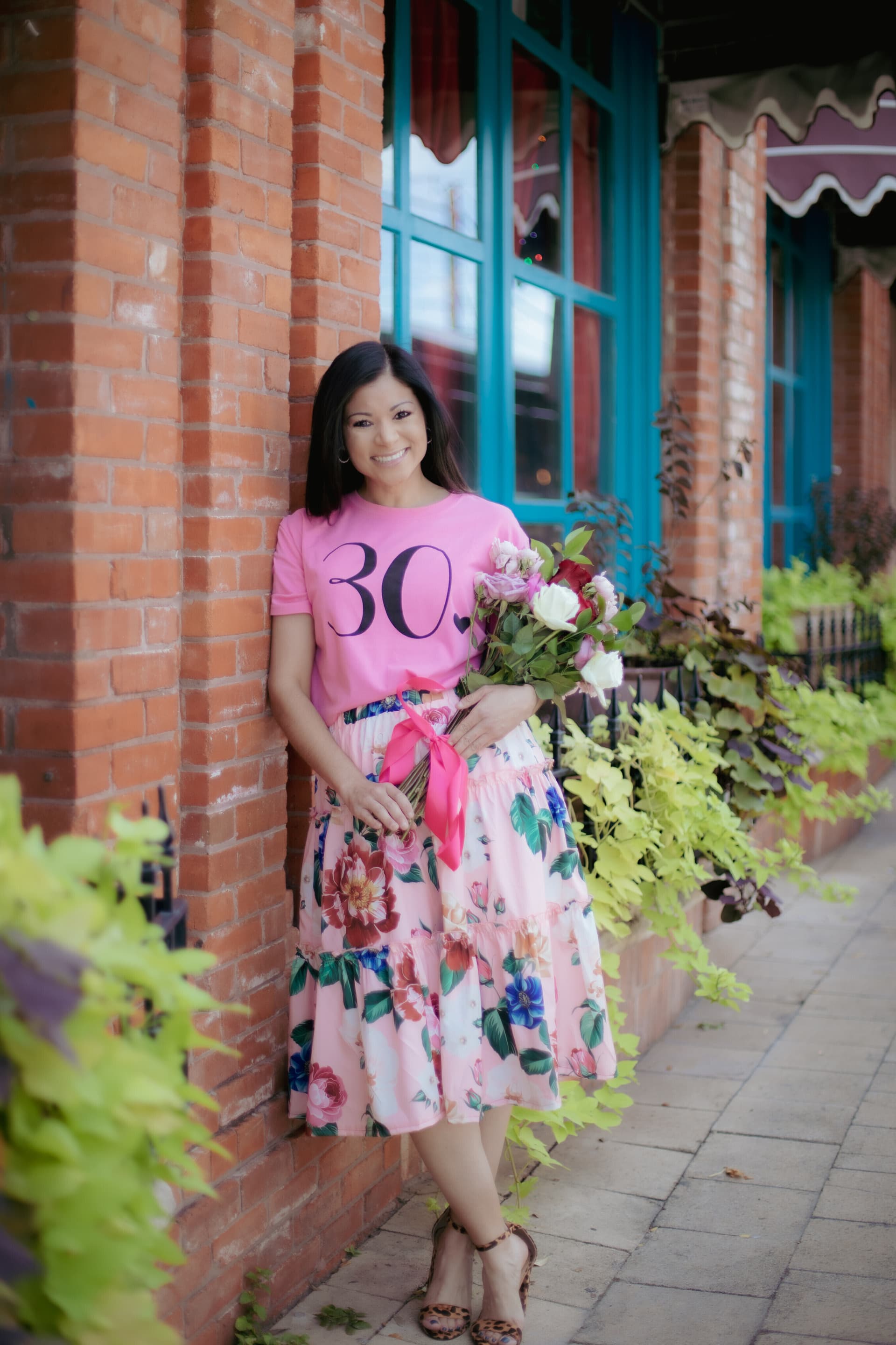 Woman in a pink '30' birthday t-shirt holding flowers while leaning against a brick storefront