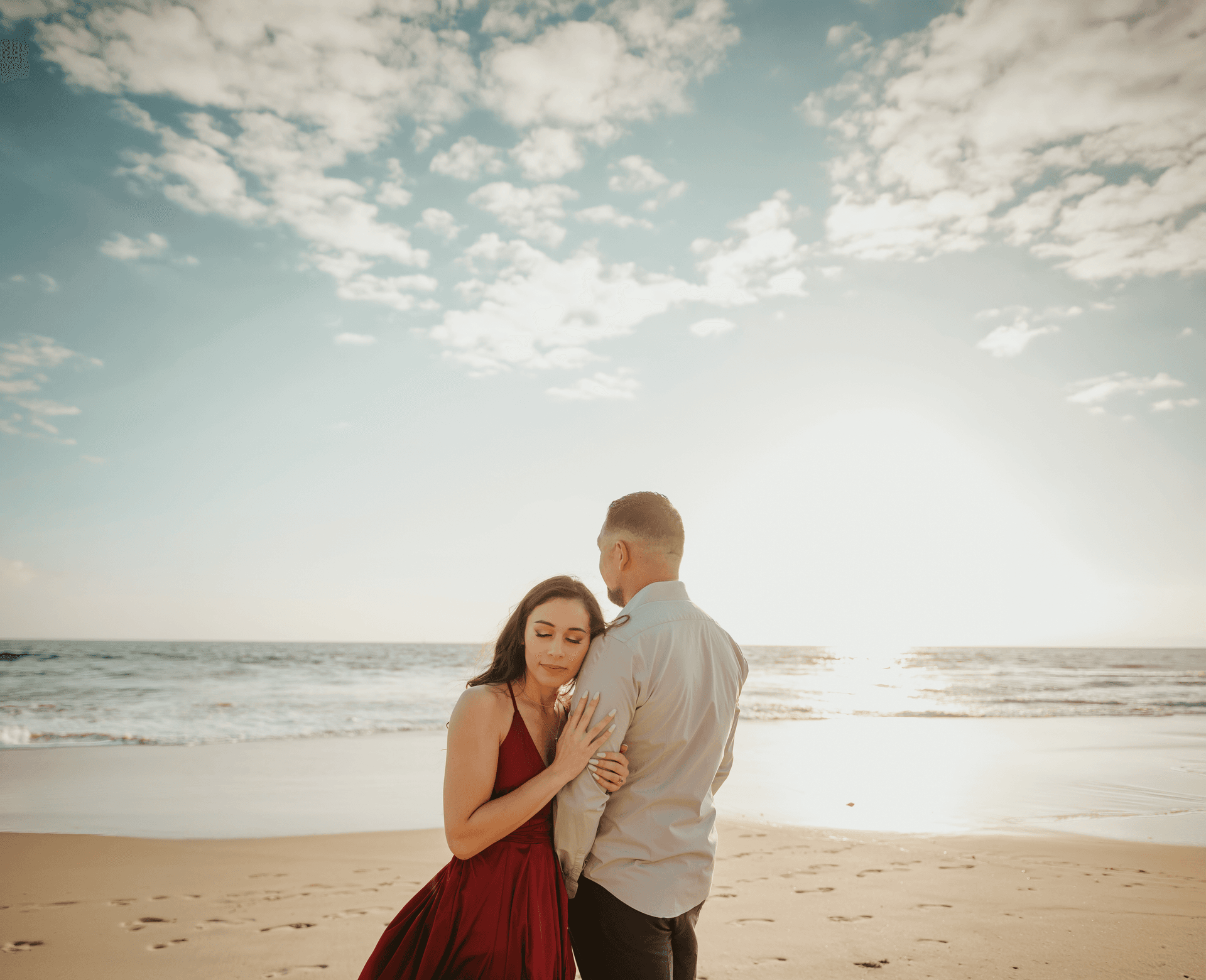 Engaged couple embracing on a sandy beach at golden hour, woman in a red dress leaning into her partner