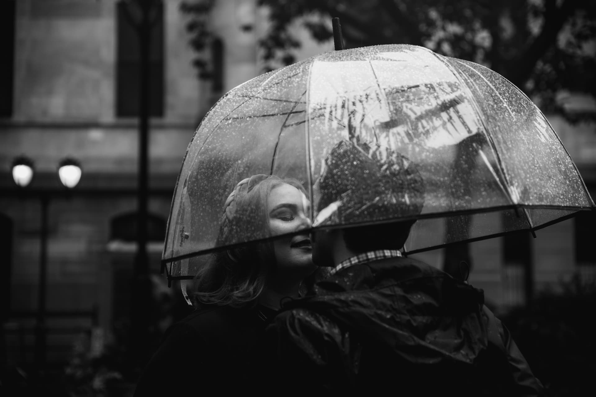 A black and white photo of two people sheltering together under a transparent umbrella in the rain