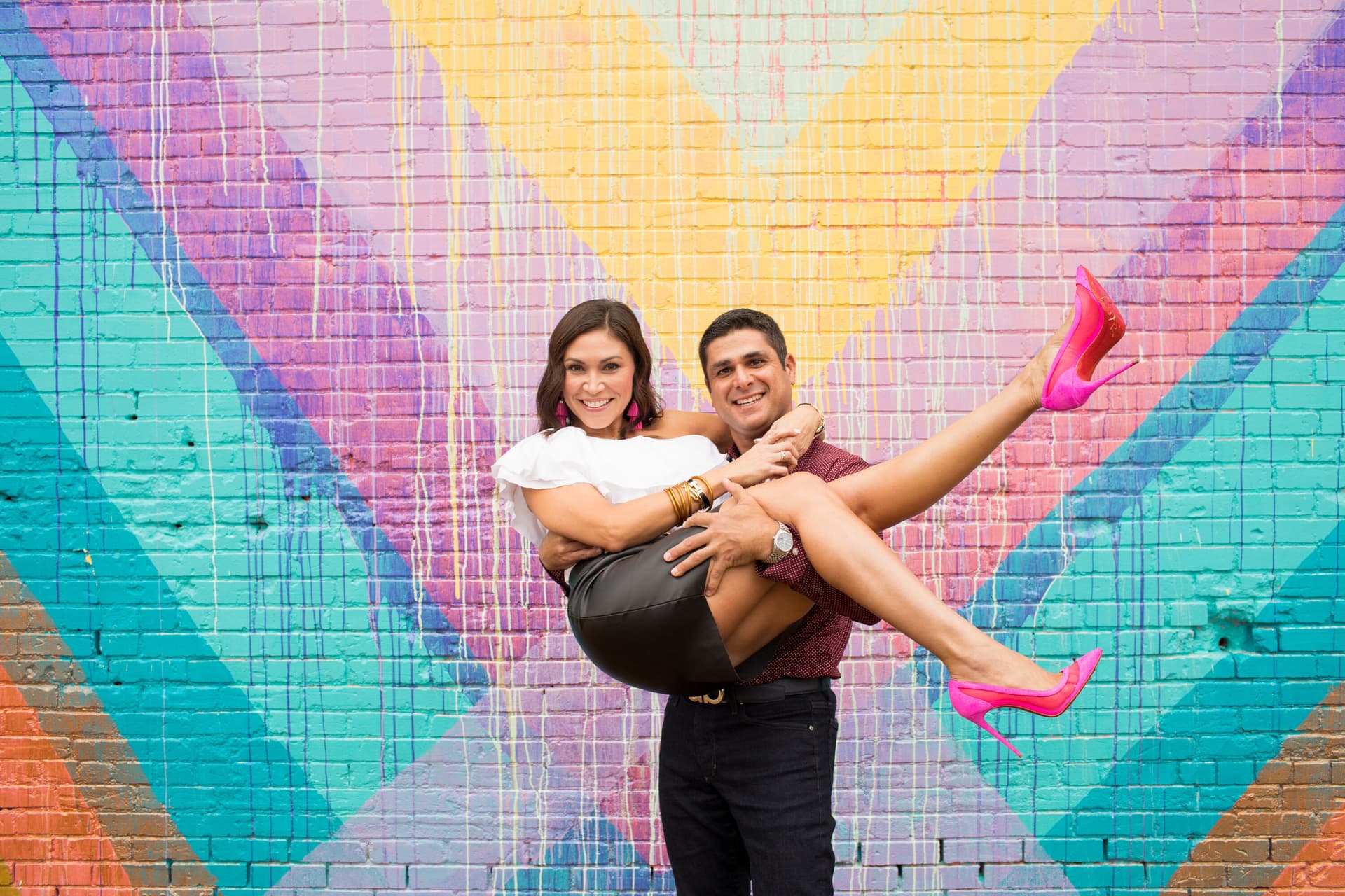 A couple posing and laughing in front of a colorful geometric mural, the woman being held up by the man