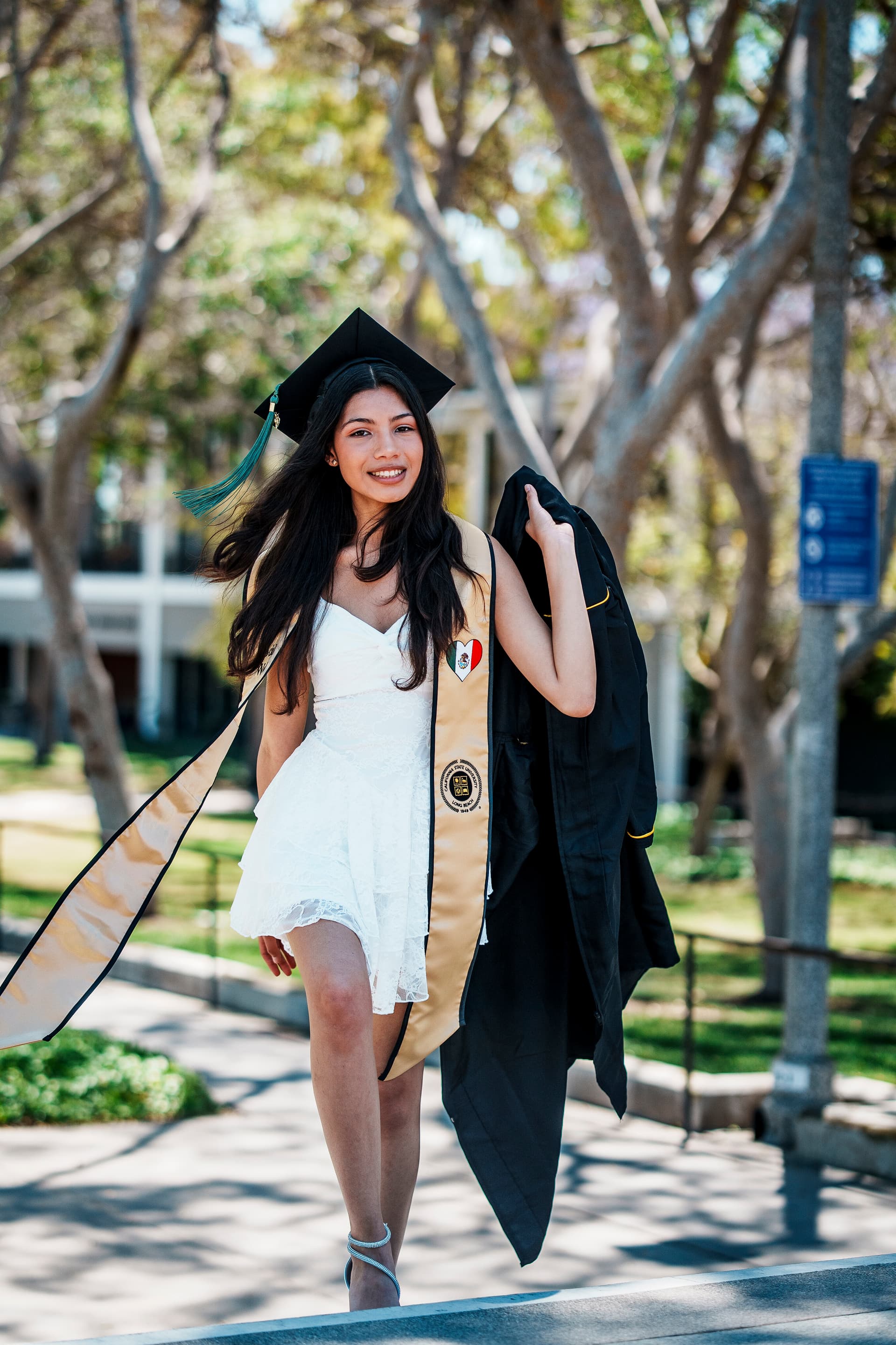 Female graduate in a white dress carrying her gown along a sunny campus path