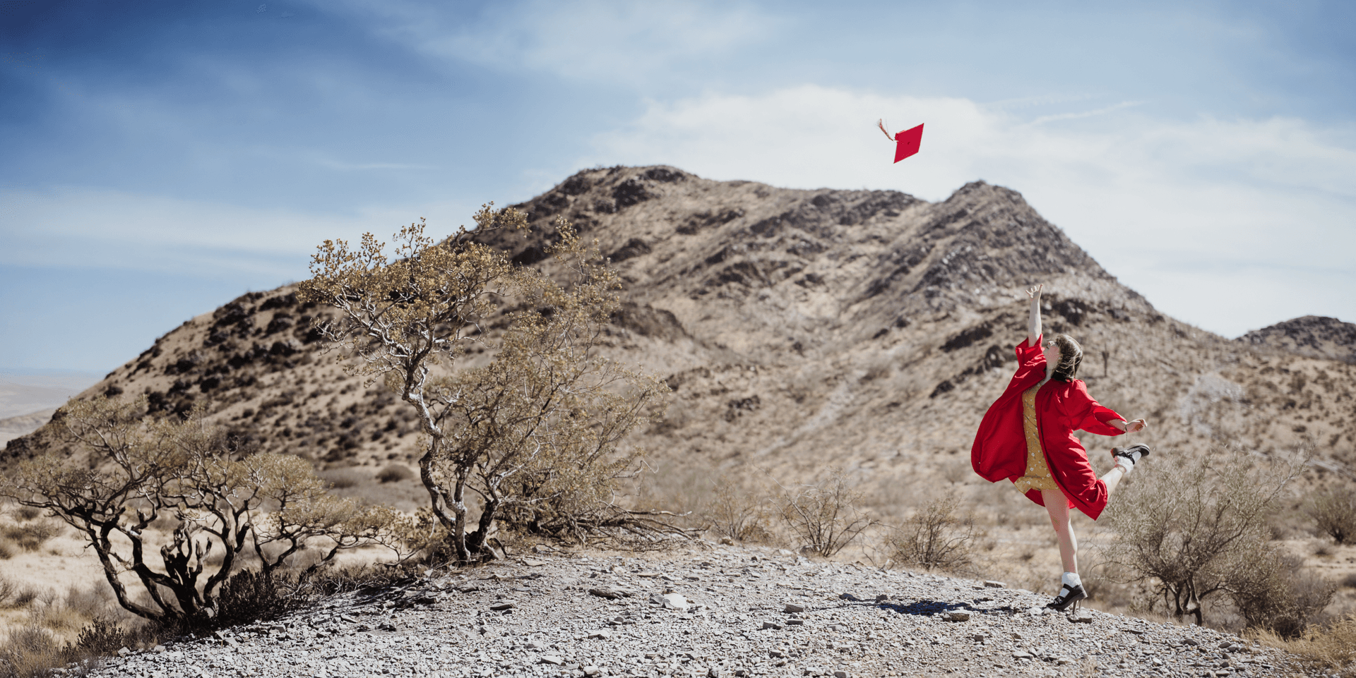 Graduate in a red dress tossing her cap in a dramatic desert landscape