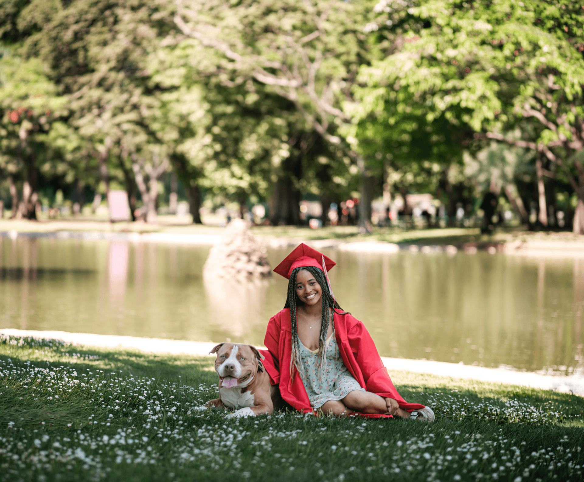 Female graduate in a red cap and gown sitting beside her bulldog in a park by a pond