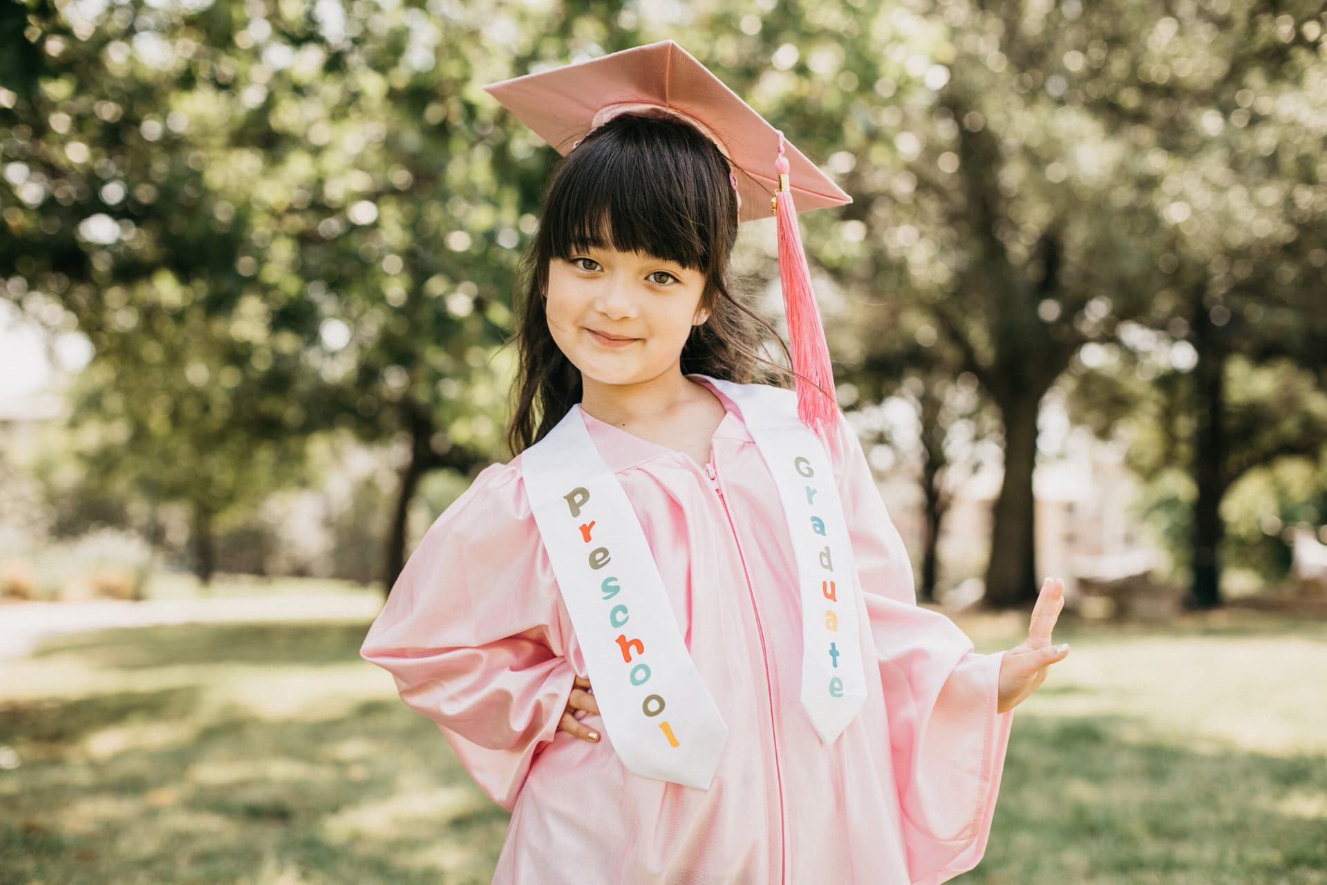 Young girl in a pink cap and gown with a preschool graduation sash