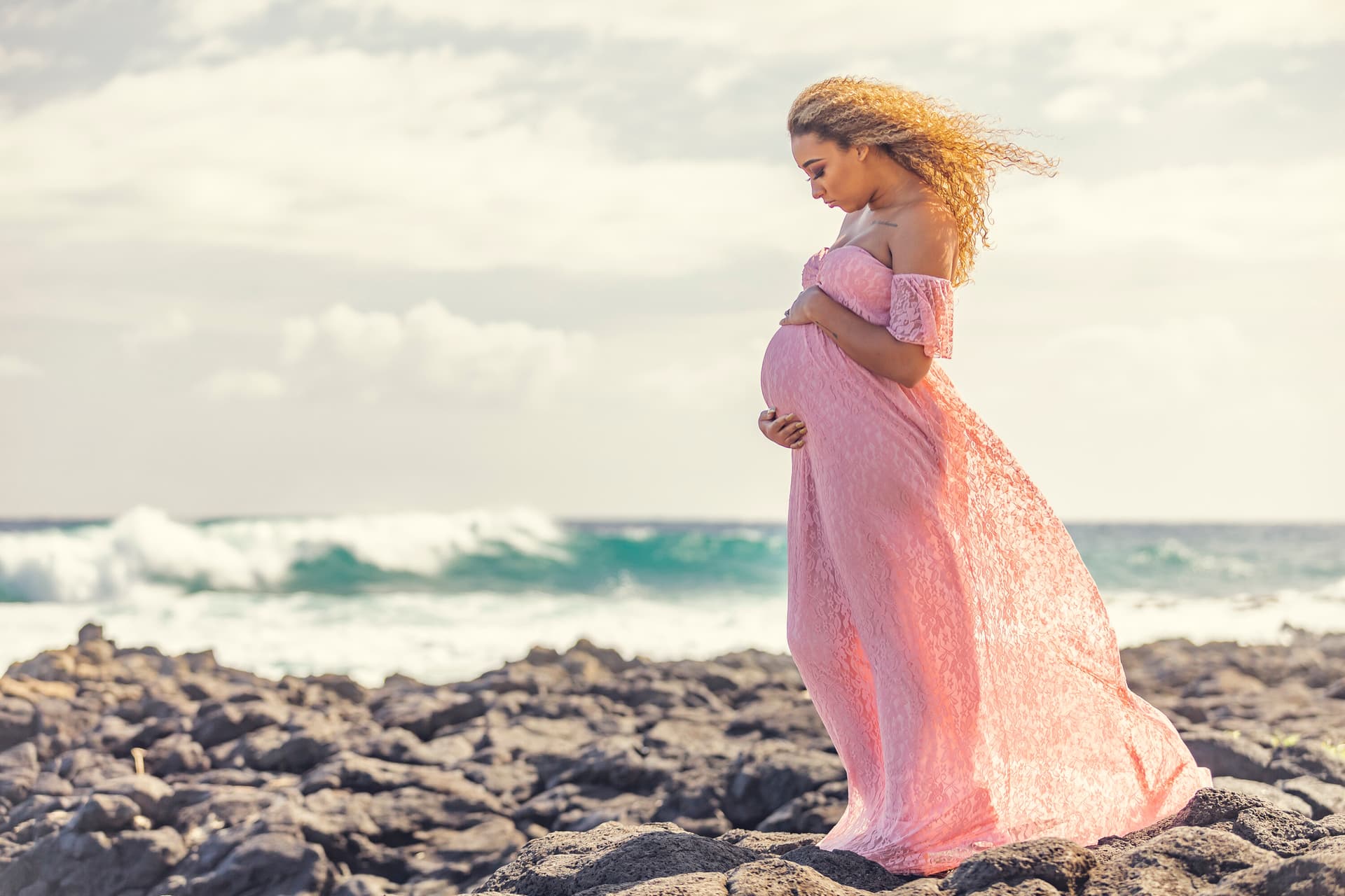 Expecting mother in a flowing pink gown standing on a rocky beach
