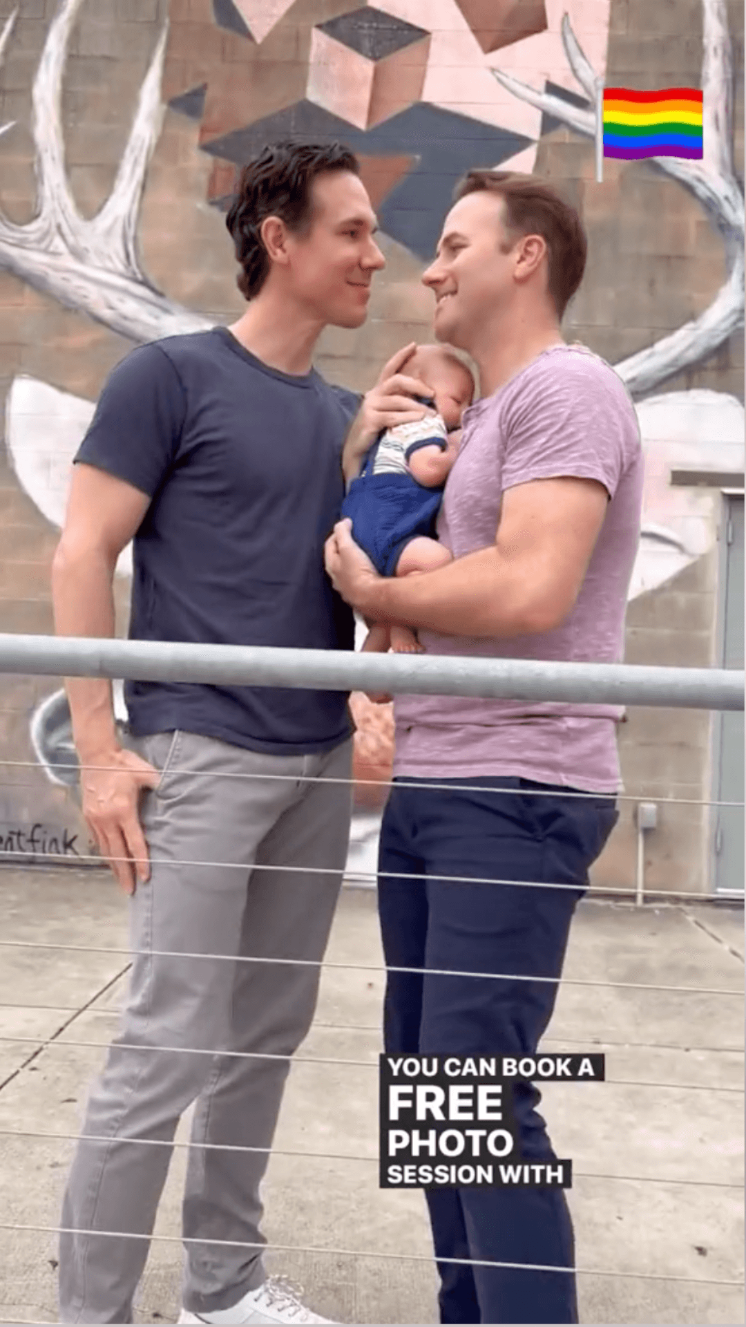 Two men smiling at each other while holding a baby, with a mural in the background.