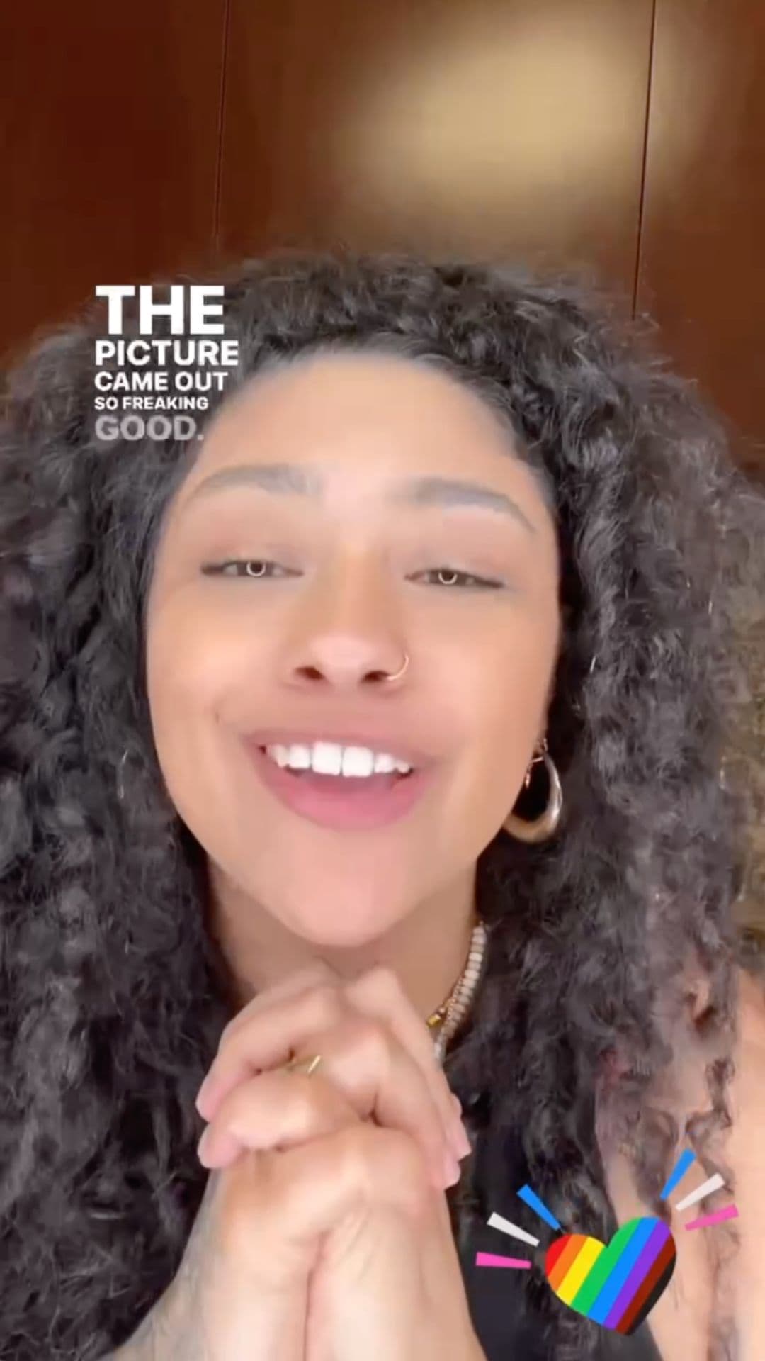 A smiling person with curly hair, a nose ring, and a small heart graphic on the cheek.