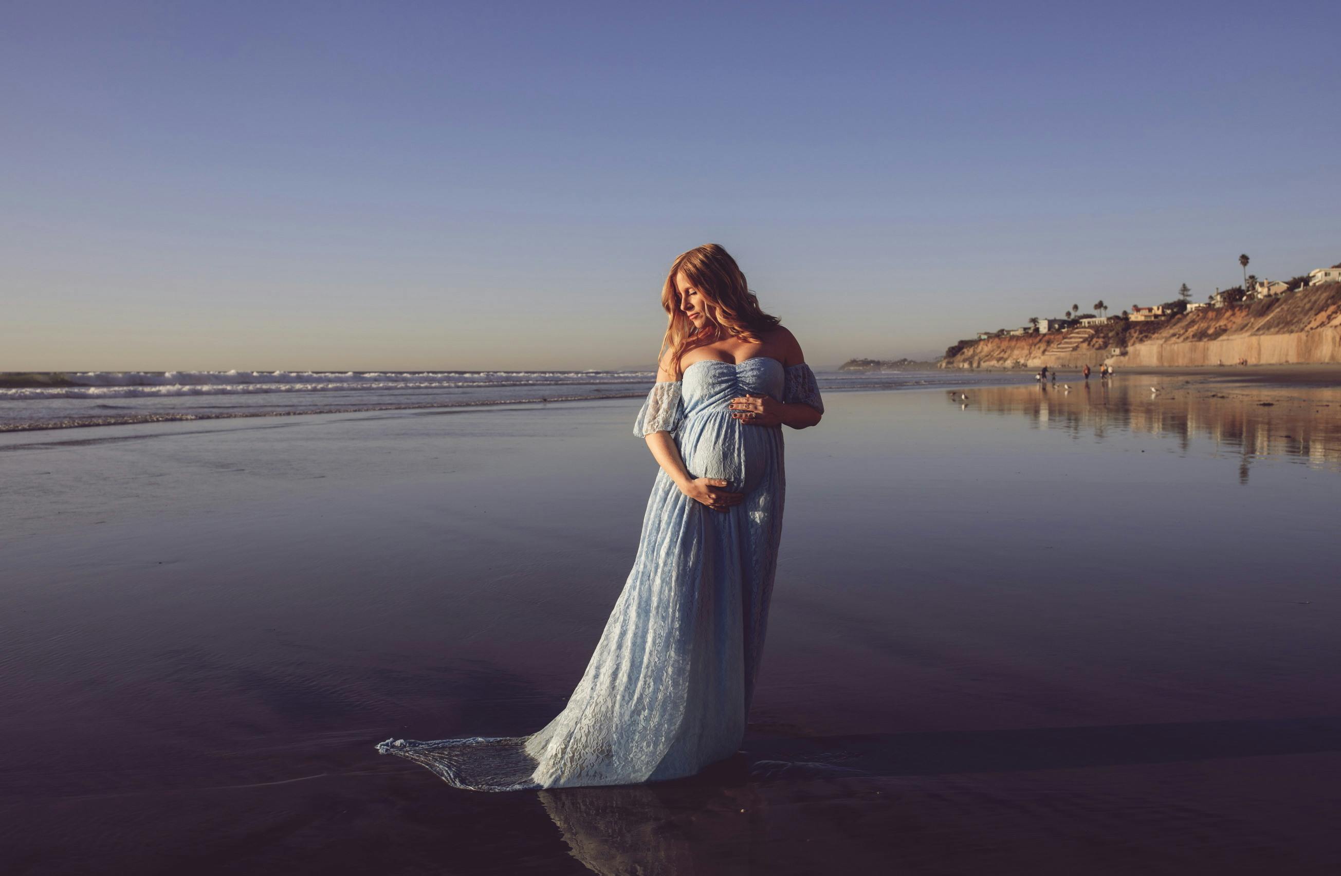 pregnant woman in a blue dress poses on the beach for her maternity photoshoot