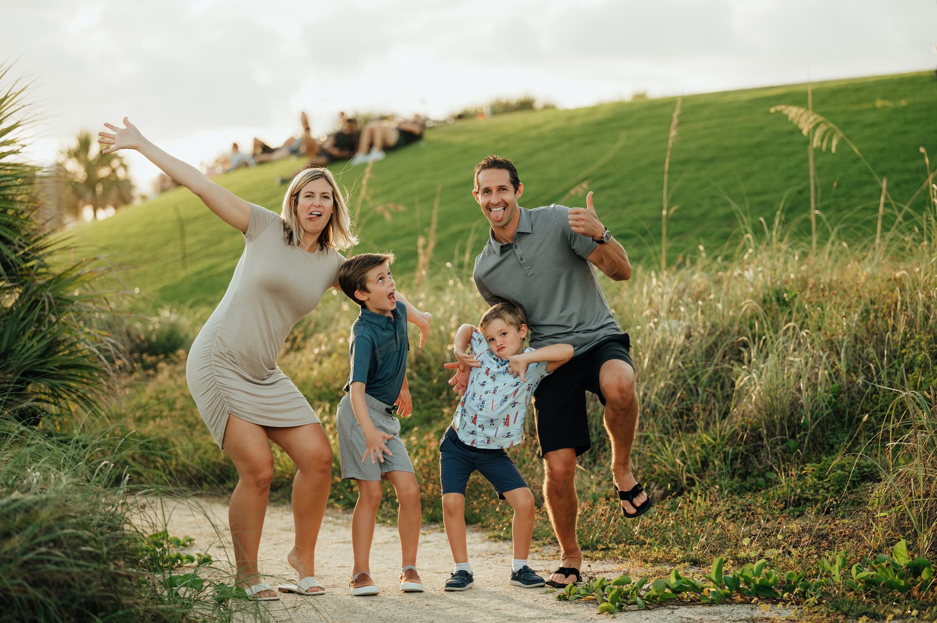 A family of four striking fun, silly poses on a sandy path surrounded by green grass and tropical plants during golden hour