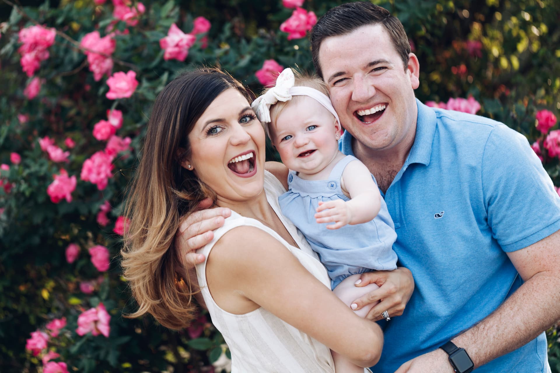 A smiling couple holds their baby girl in front of a bright pink flowering bush