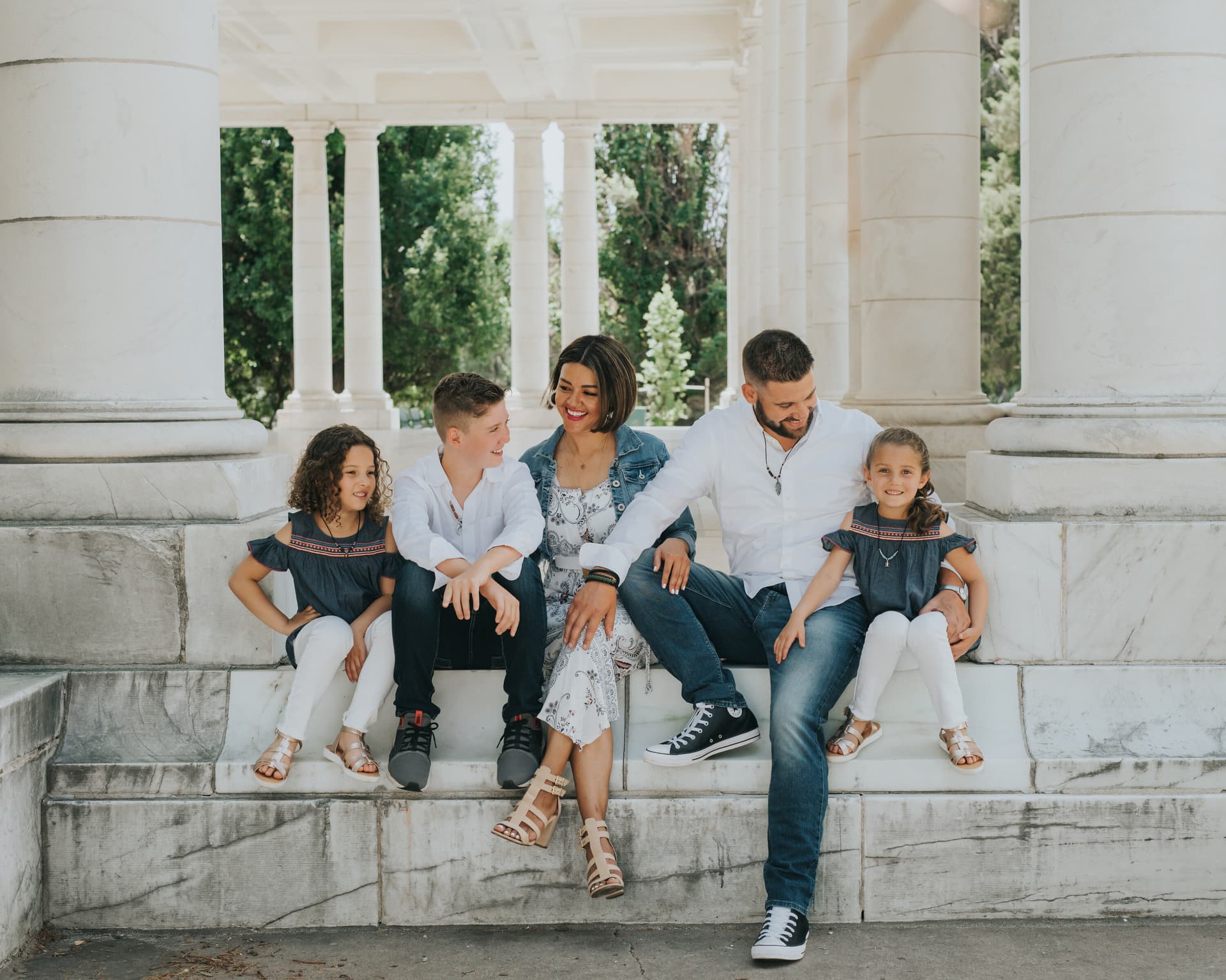A family of five sits together on stone steps, laughing and interacting warmly in an outdoor setting with columns and trees