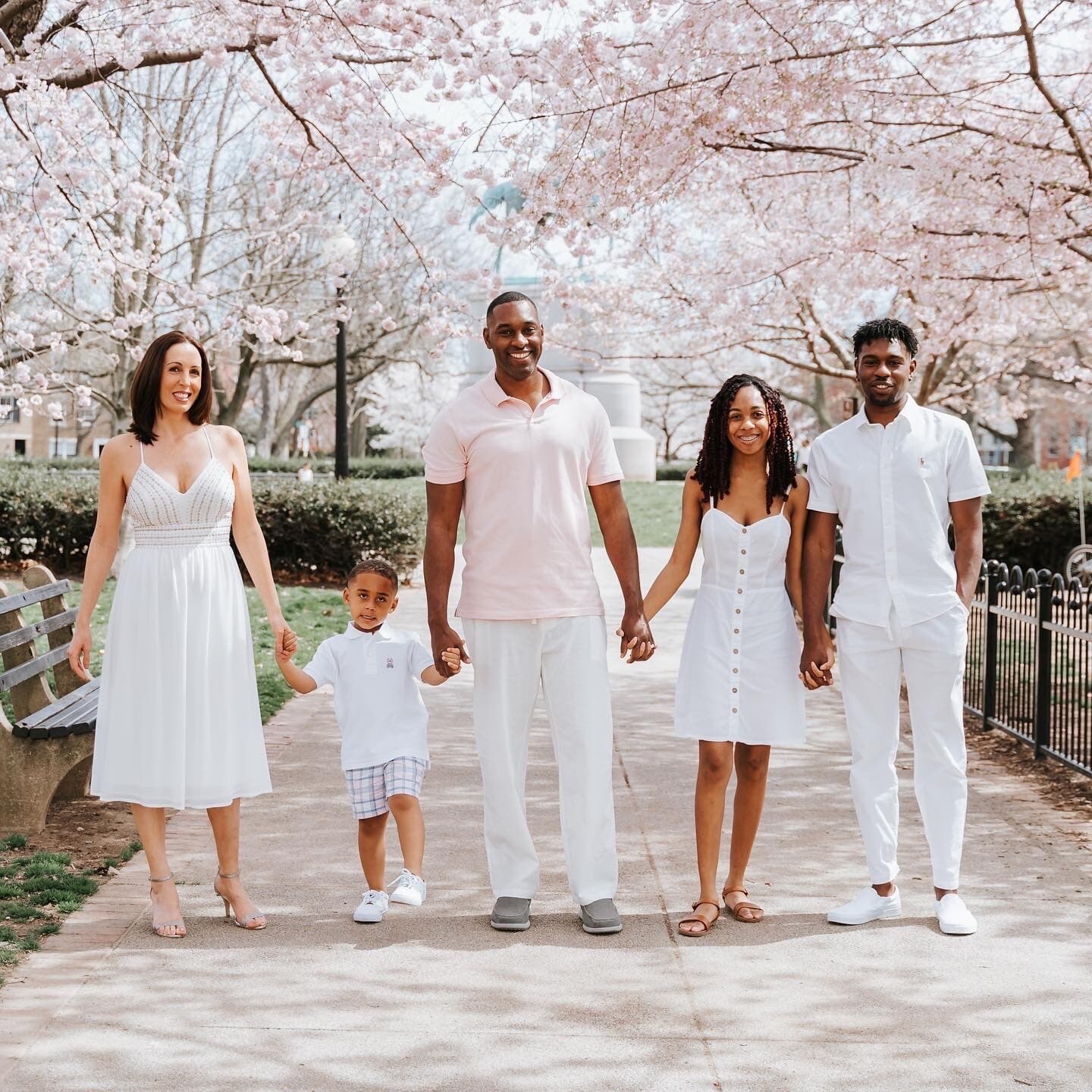 A family of four dressed in white walk hand-in-hand along a path lined with blooming cherry blossom trees in spring