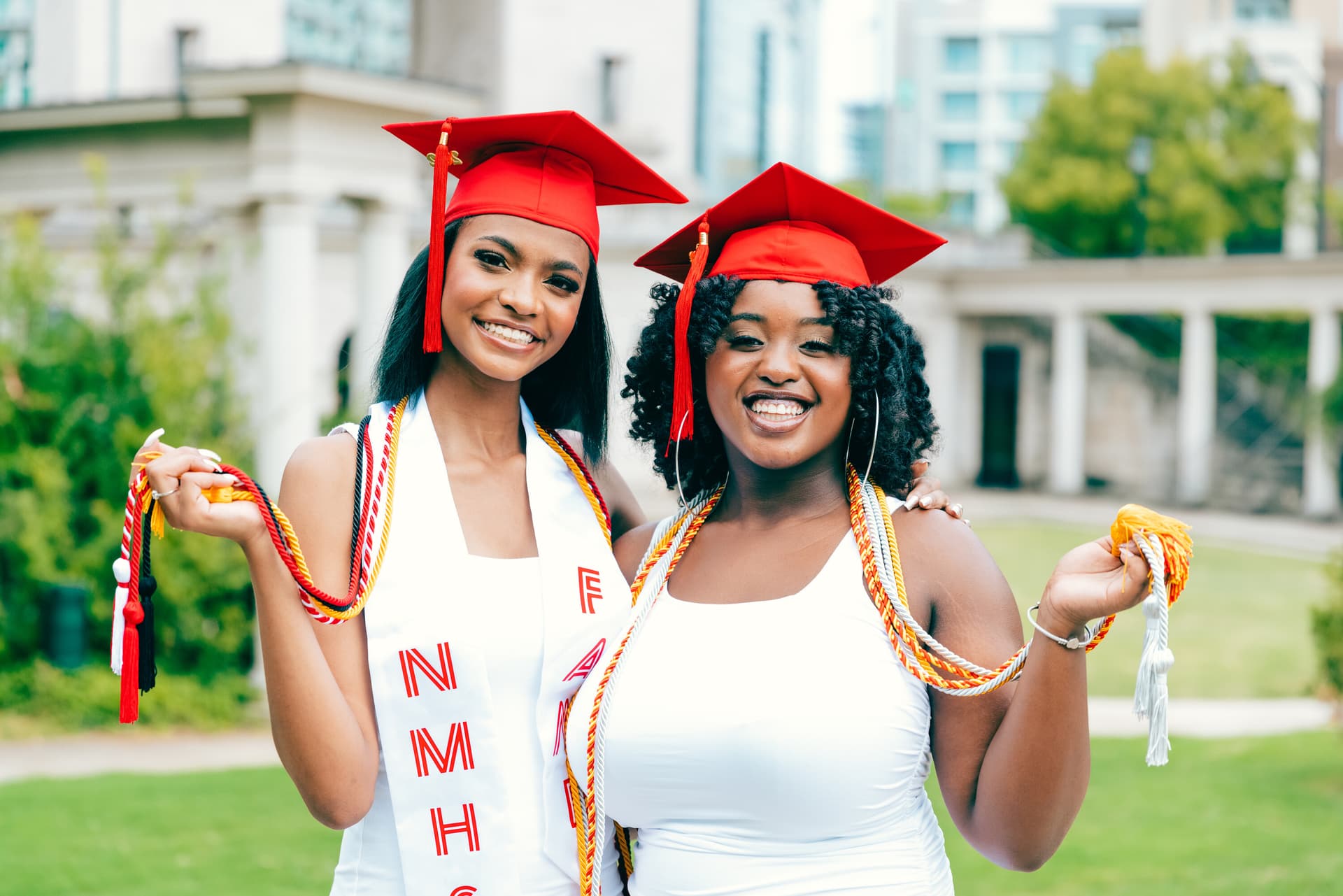 Two young women in red graduation caps and gowns smile proudly outdoors, holding diplomas and wearing colorful graduation stoles
