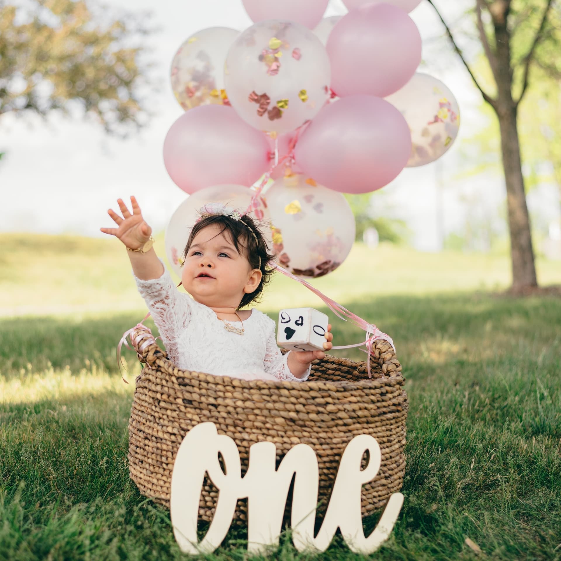 A baby sits inside a wicker basket surrounded by pink balloons and confetti, reaching upward joyfully, with an "One" sign in front