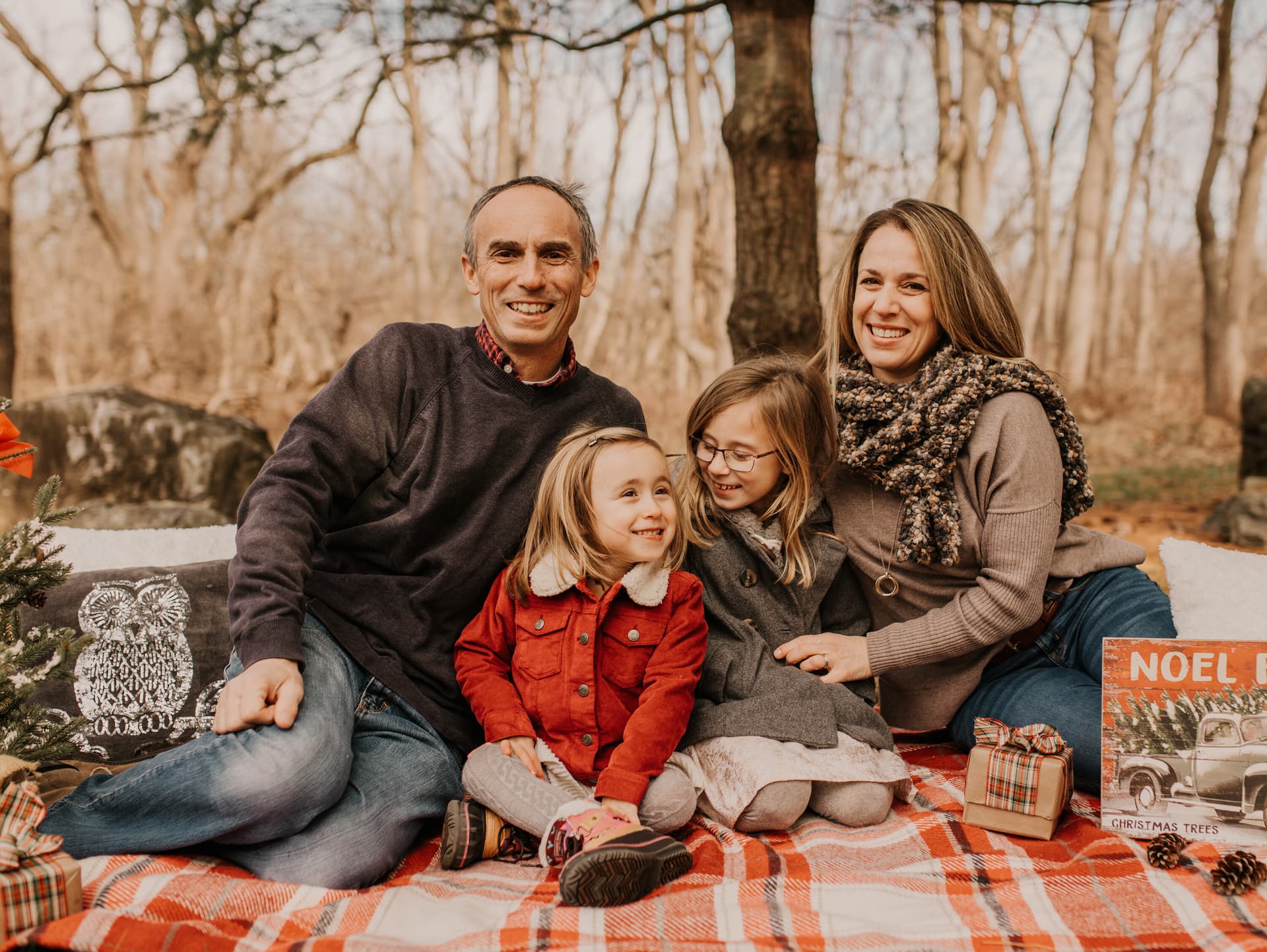 A family of five huddles closely together on a plaid blanket in a park with bare autumn trees, smiling warmly at the camera
