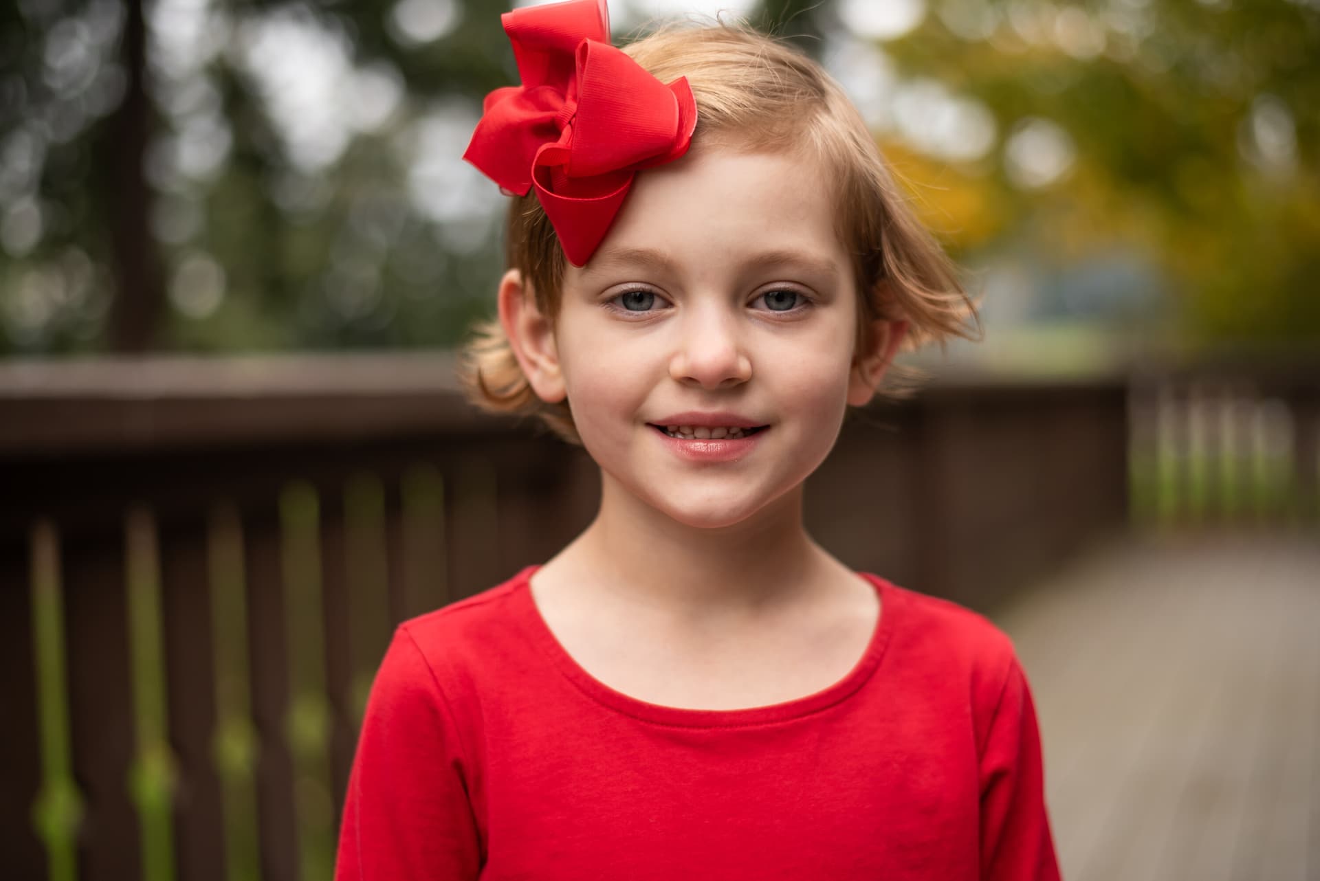 A young girl with a large red bow in her hair smiles softly at the camera against a blurred outdoor background