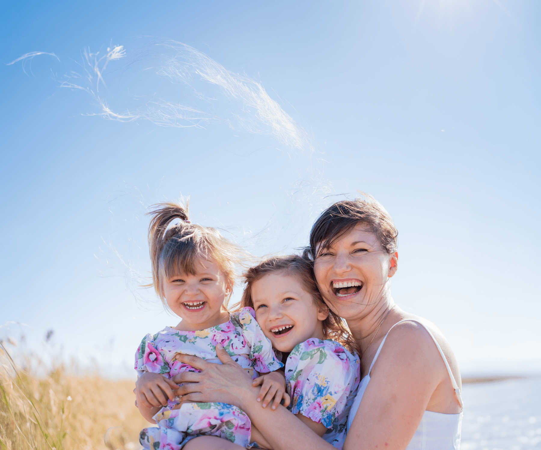 Mother laughing joyfully while holding her two young daughters in matching floral dresses outdoors on a sunny day