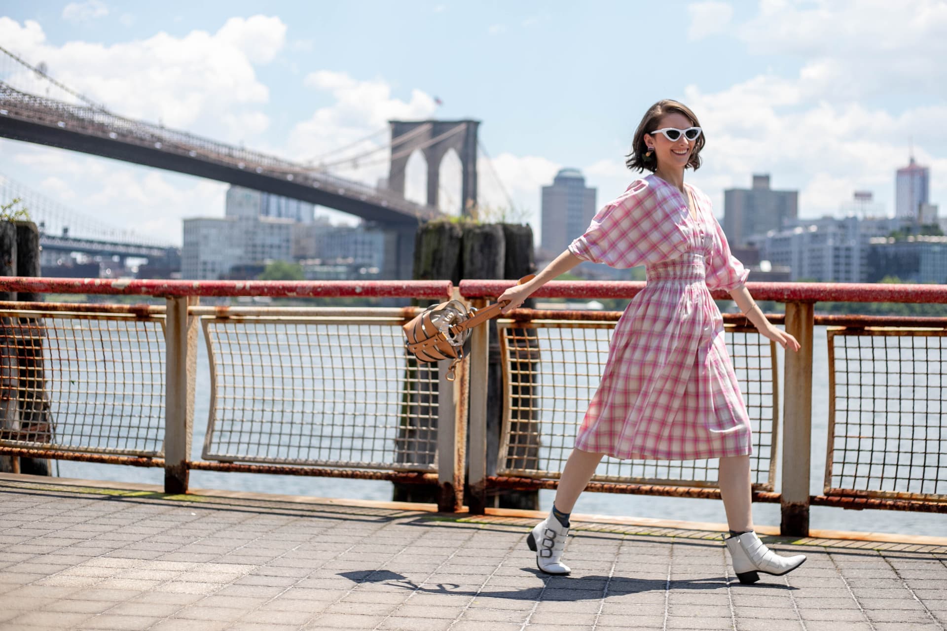 A woman in a pink gingham dress posing playfully on a waterfront boardwalk with the Brooklyn Bridge in the background