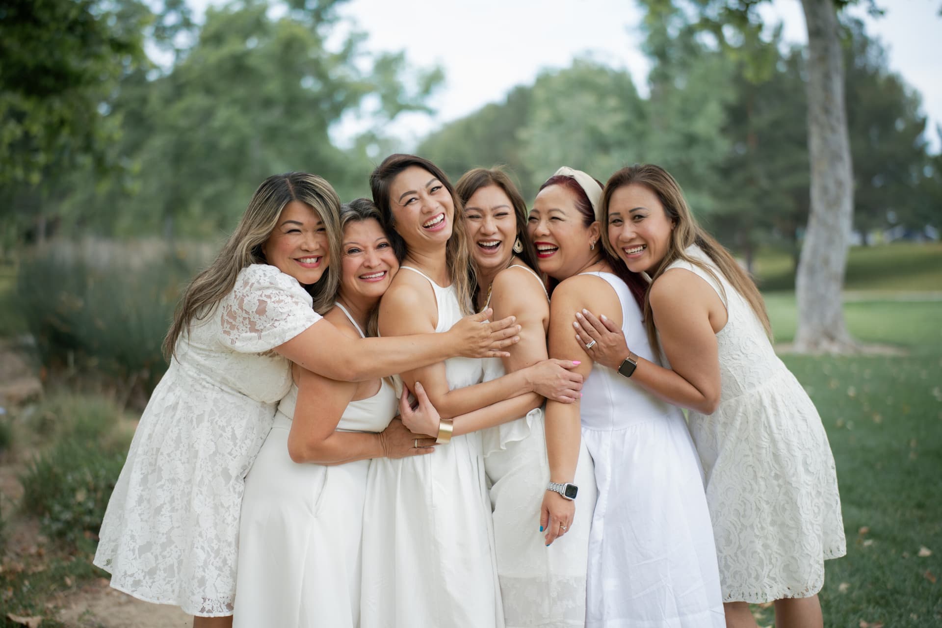A group of six women in white dresses laughing and hugging each other in a lush green park setting
