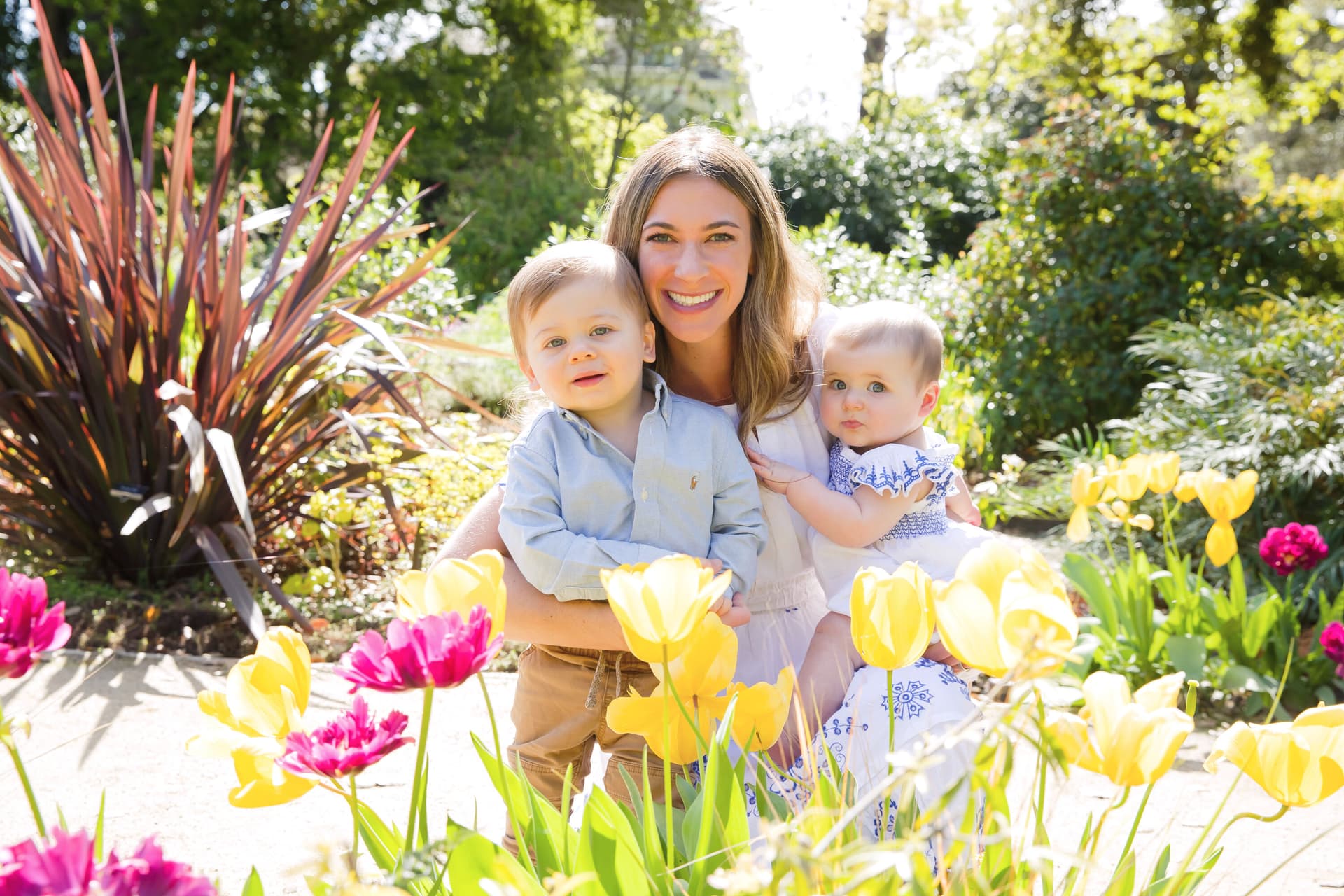 A woman sitting outdoors in a garden surrounded by colorful tulips, holding two young children on her lap