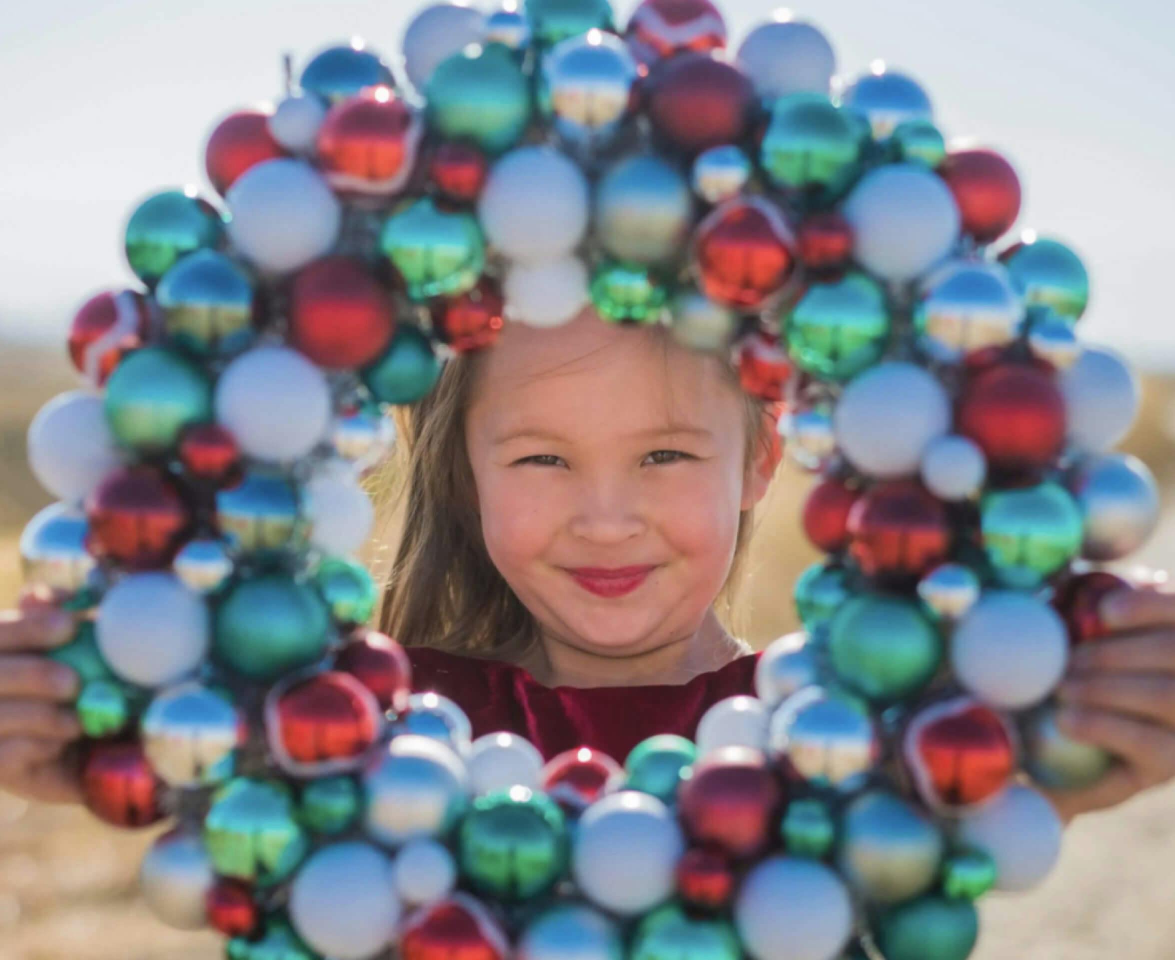 girl looking at the camera through a wreath made of christmas decorations