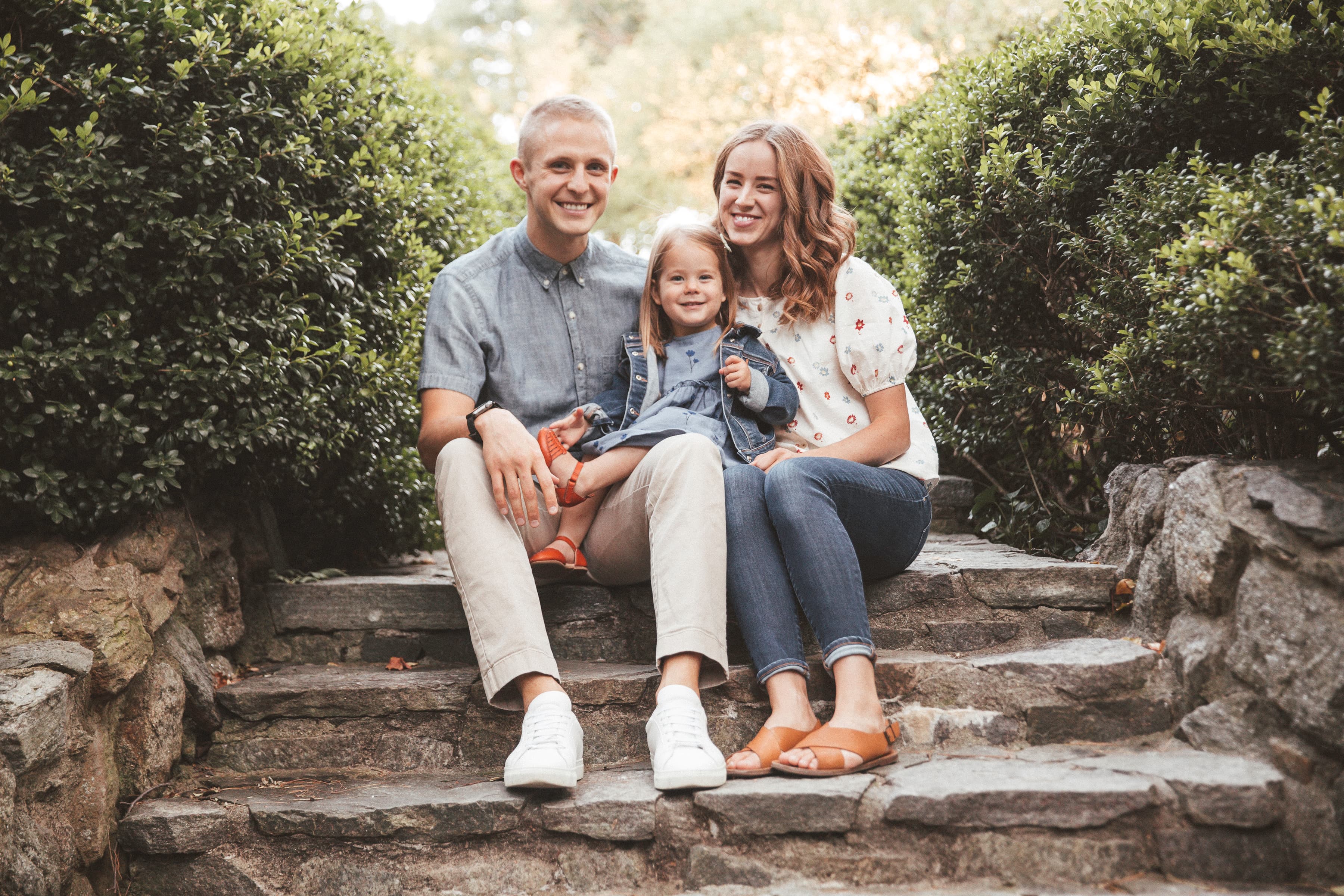 A family of three smiling and sitting on outdoor steps surrounded by greenery.