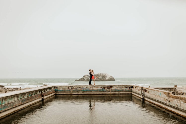 A couple kissing on a pier with the ocean and rock formations in the background