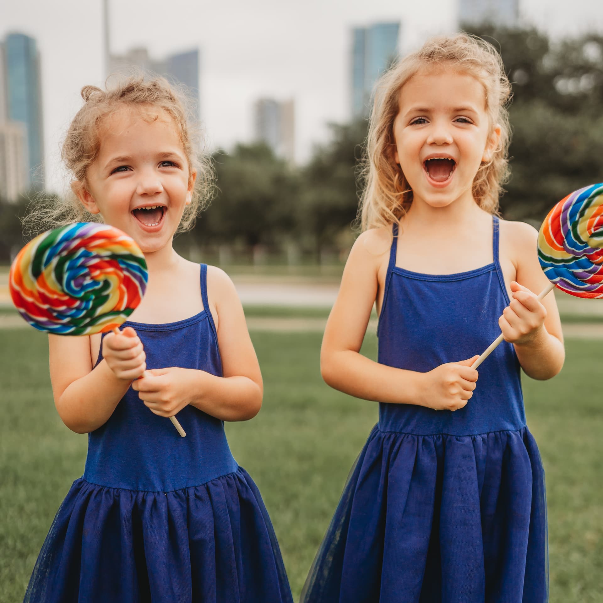 Two joyful girls in blue dresses holding large lollipops in a park.