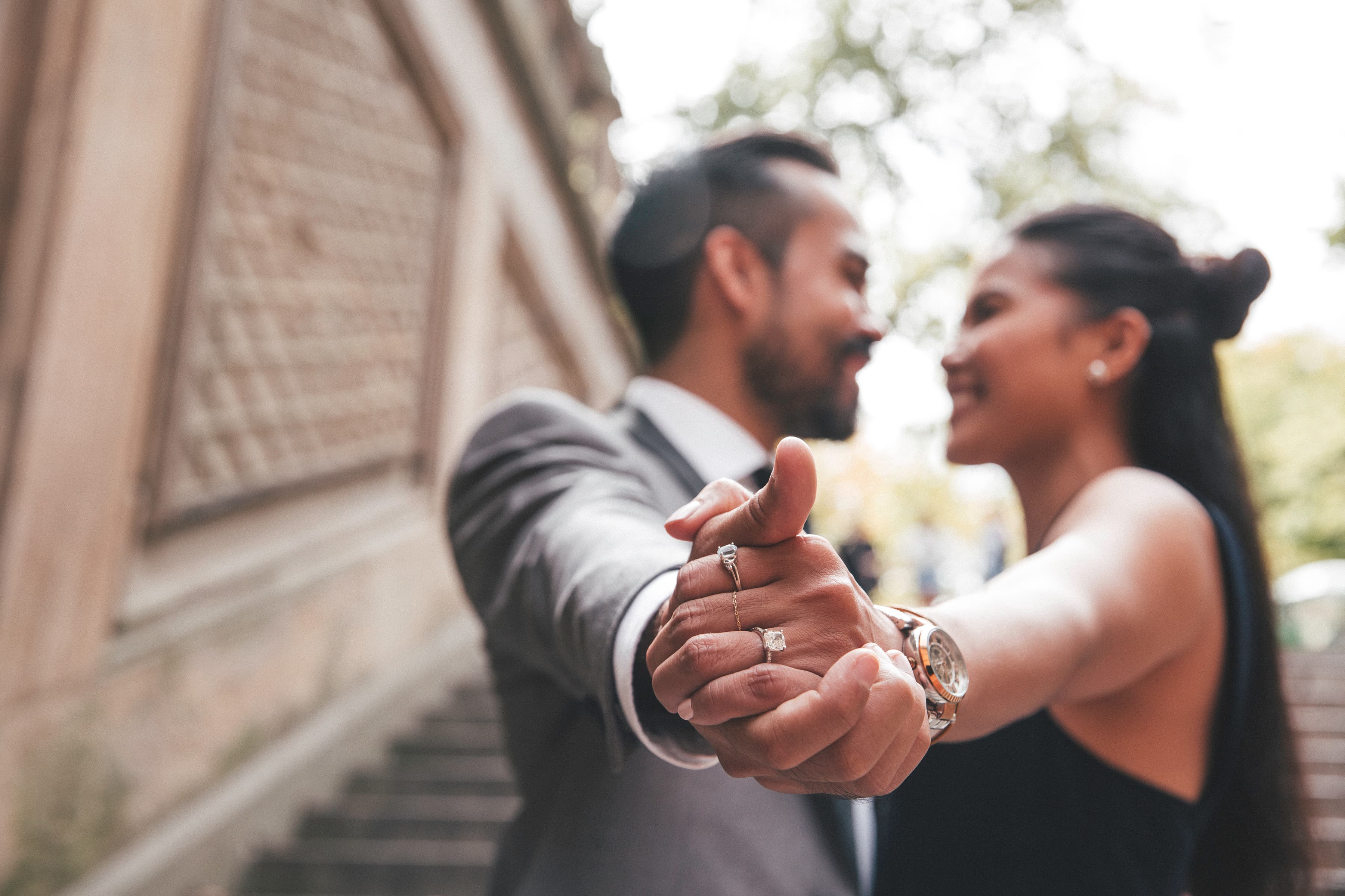 A couple holding hands and smiling at each other, showing an engagement ring.