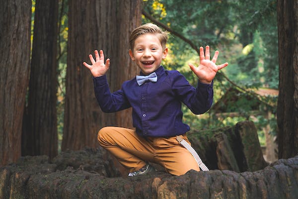 A boy in formal attire showing his hands and smiling in a forest.