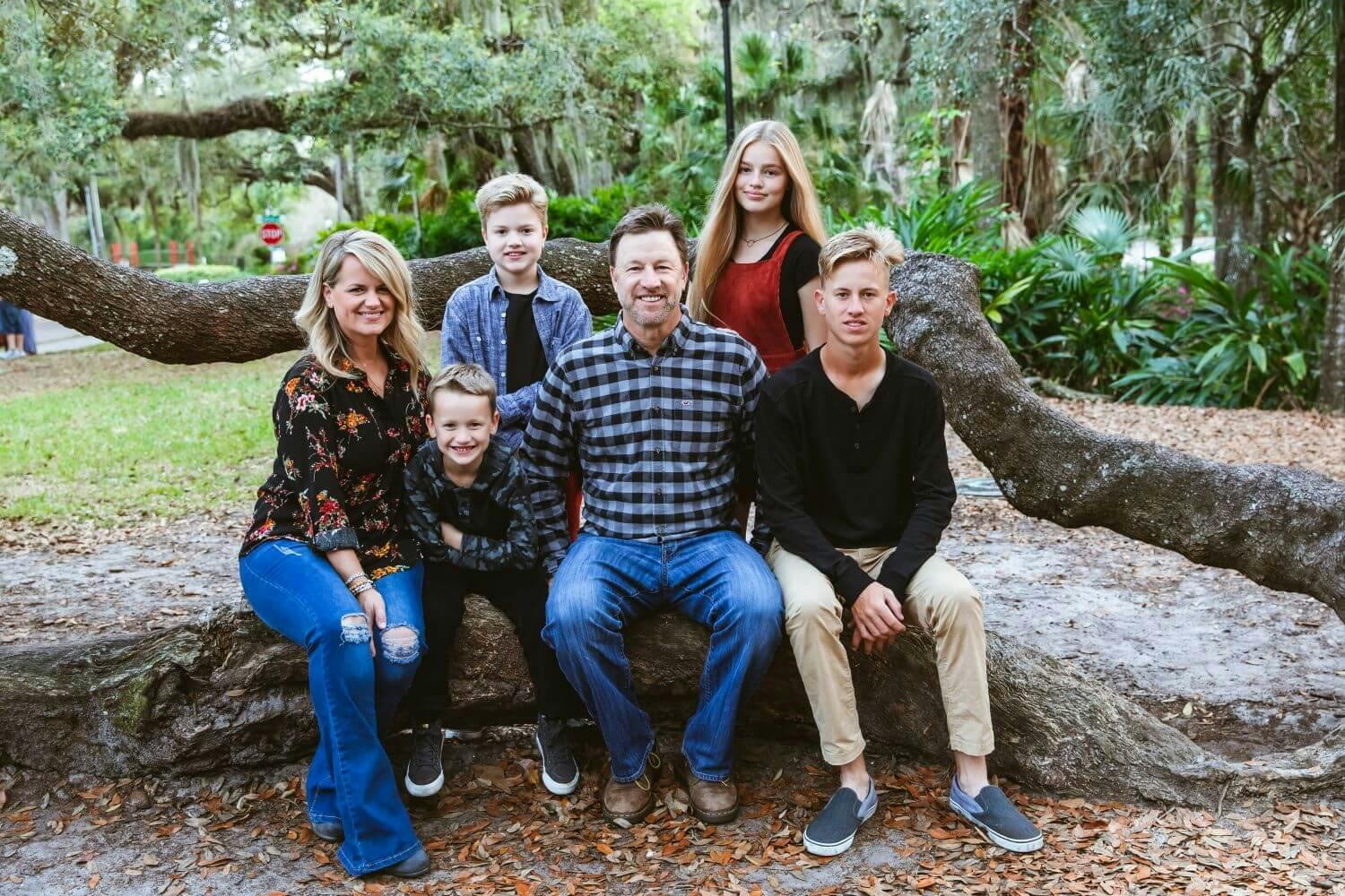A family of six smiling and sitting on a large tree branch in a park setting.