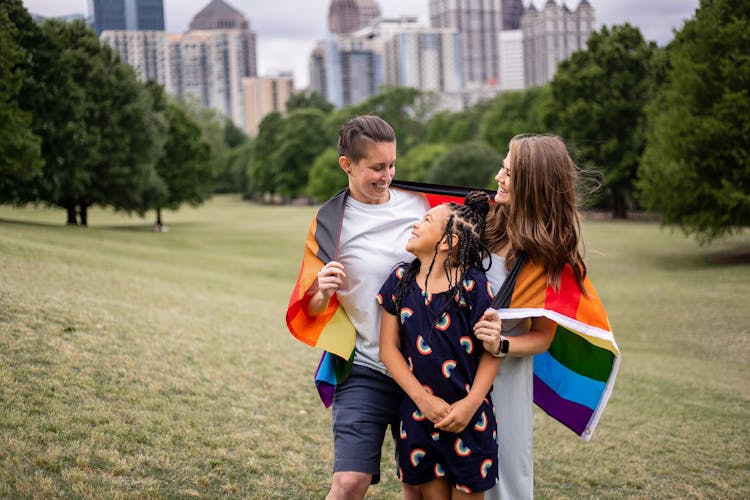 Three people smiling and walking in a park with one wearing a rainbow flag.