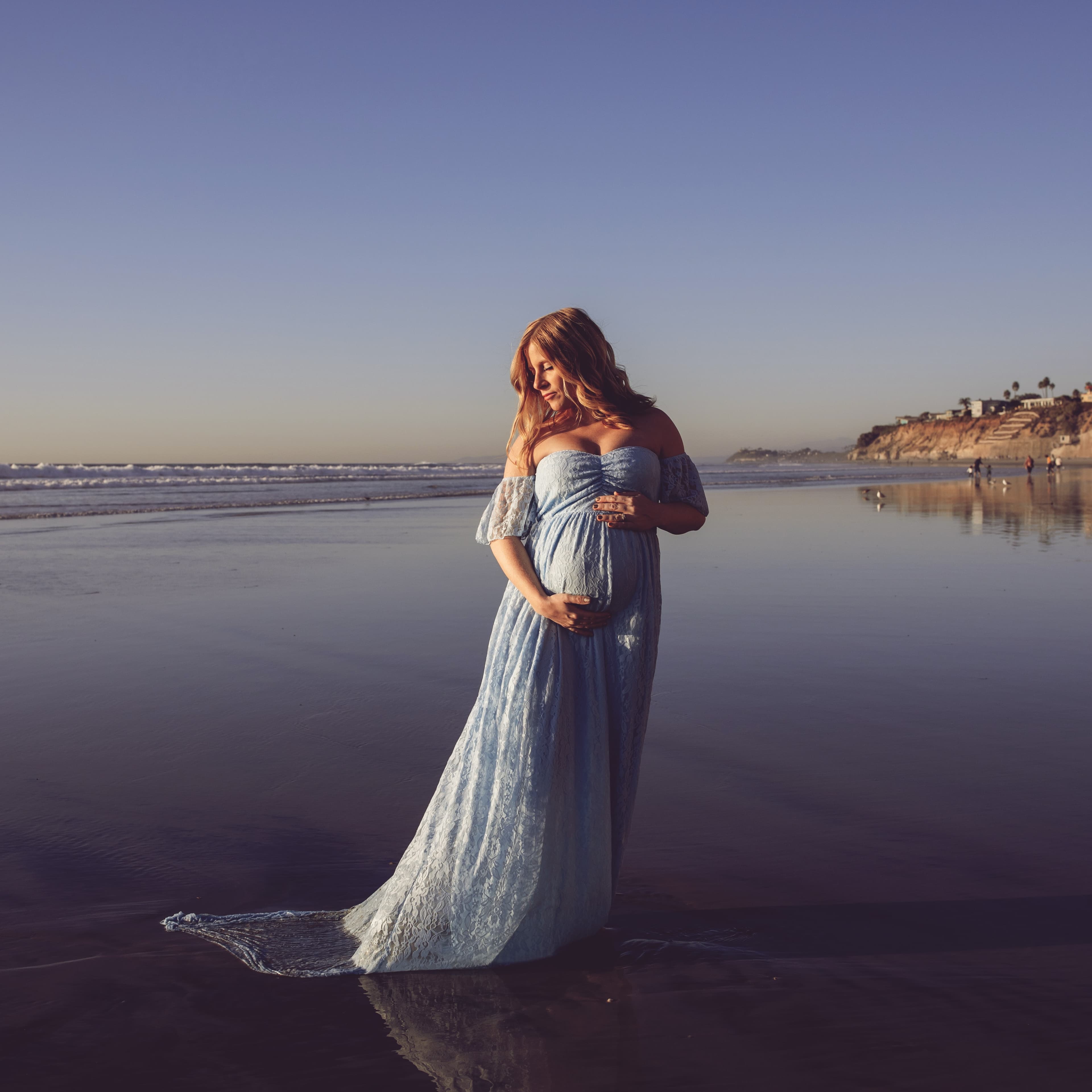 Pregnant woman in a blue dress standing on the beach at sunset.