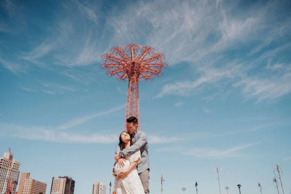 Couple embracing in front of a sky-blue backdrop with a tall metal structure.