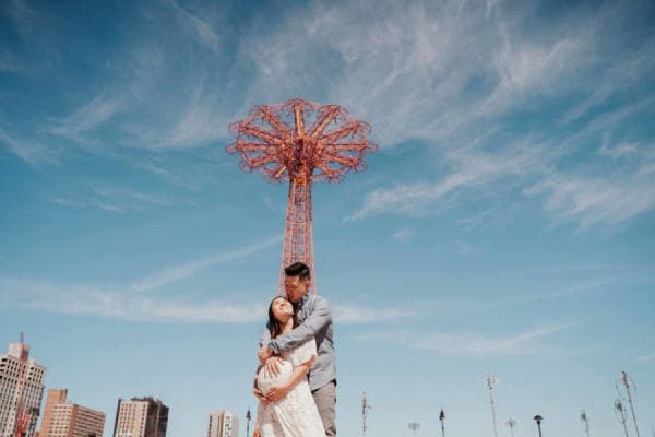 Couple embracing in front of a sky-blue backdrop with a tall metal structure.