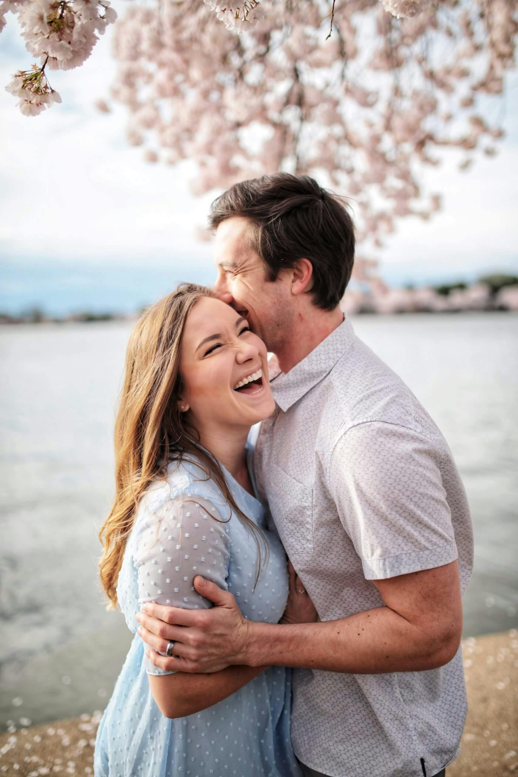 A man and woman embracing and laughing together with cherry blossoms in the background.