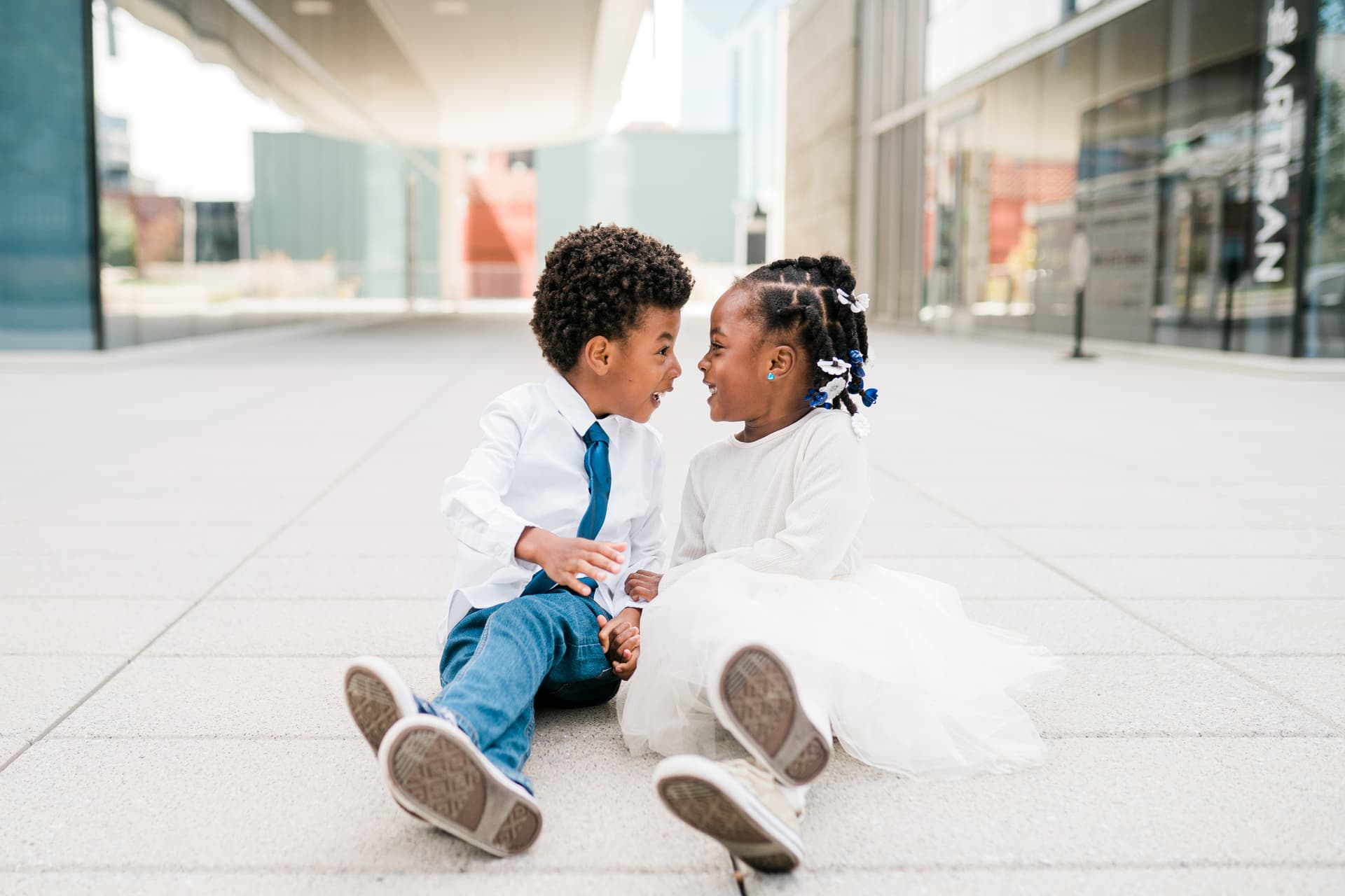 Two young children sitting on a sidewalk, smiling and looking at each other.