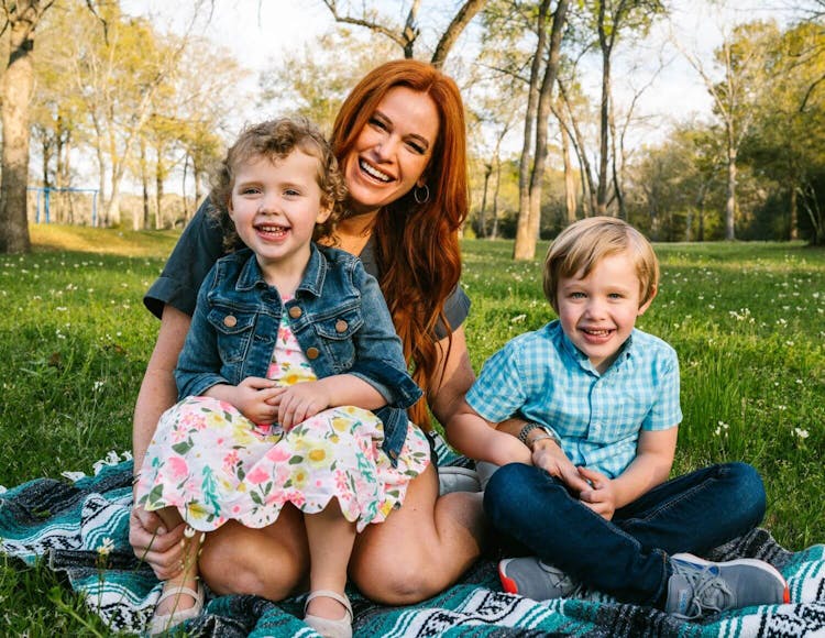 A woman seated on a blanket in a park with two young children smiling at the camera.