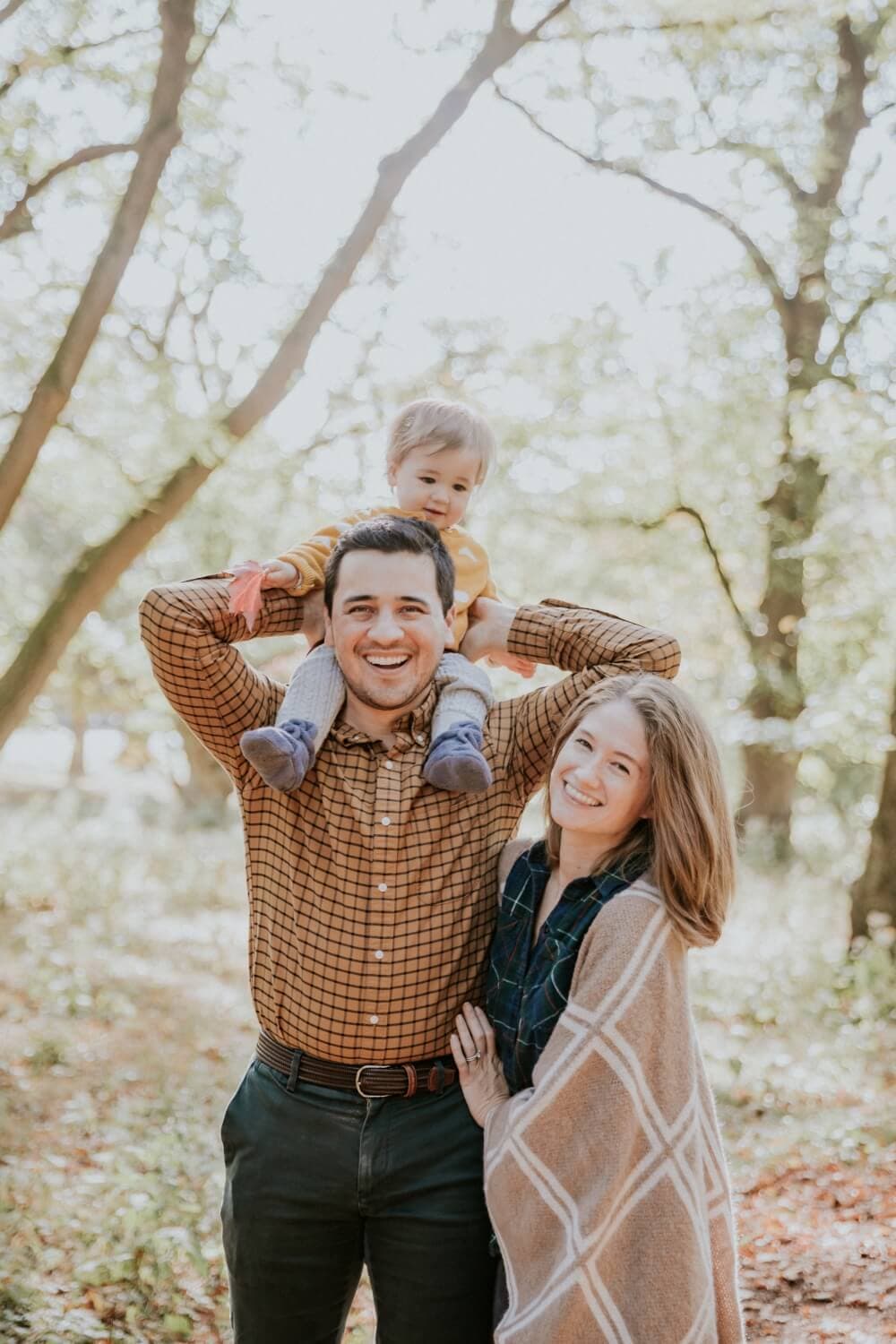 A family with a child on the father's shoulders, all smiling in a forest setting.