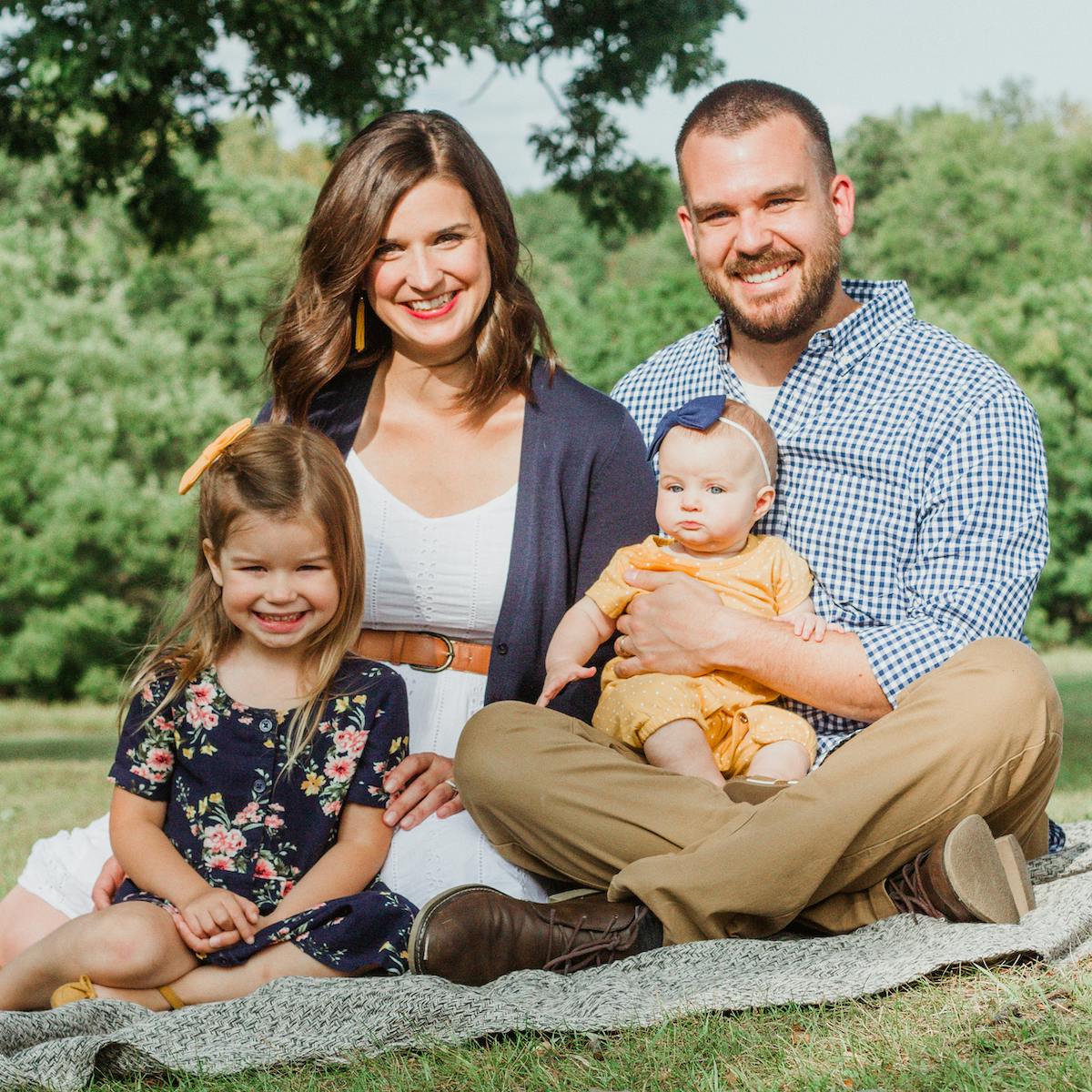 A family portrait of two adults and two children smiling and sitting on a blanket outdoors.