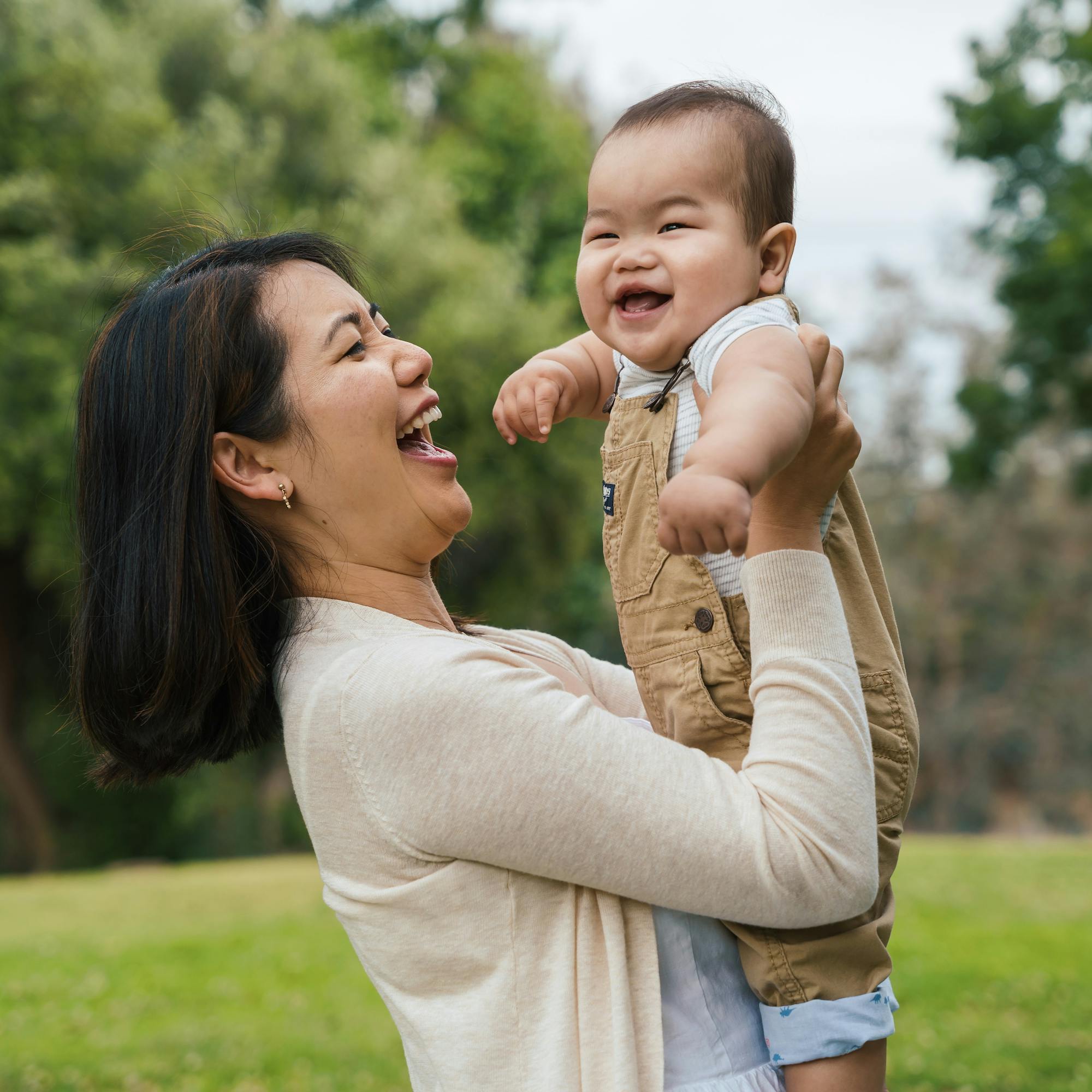 A joyful mother lifting her laughing baby in the air at a park.