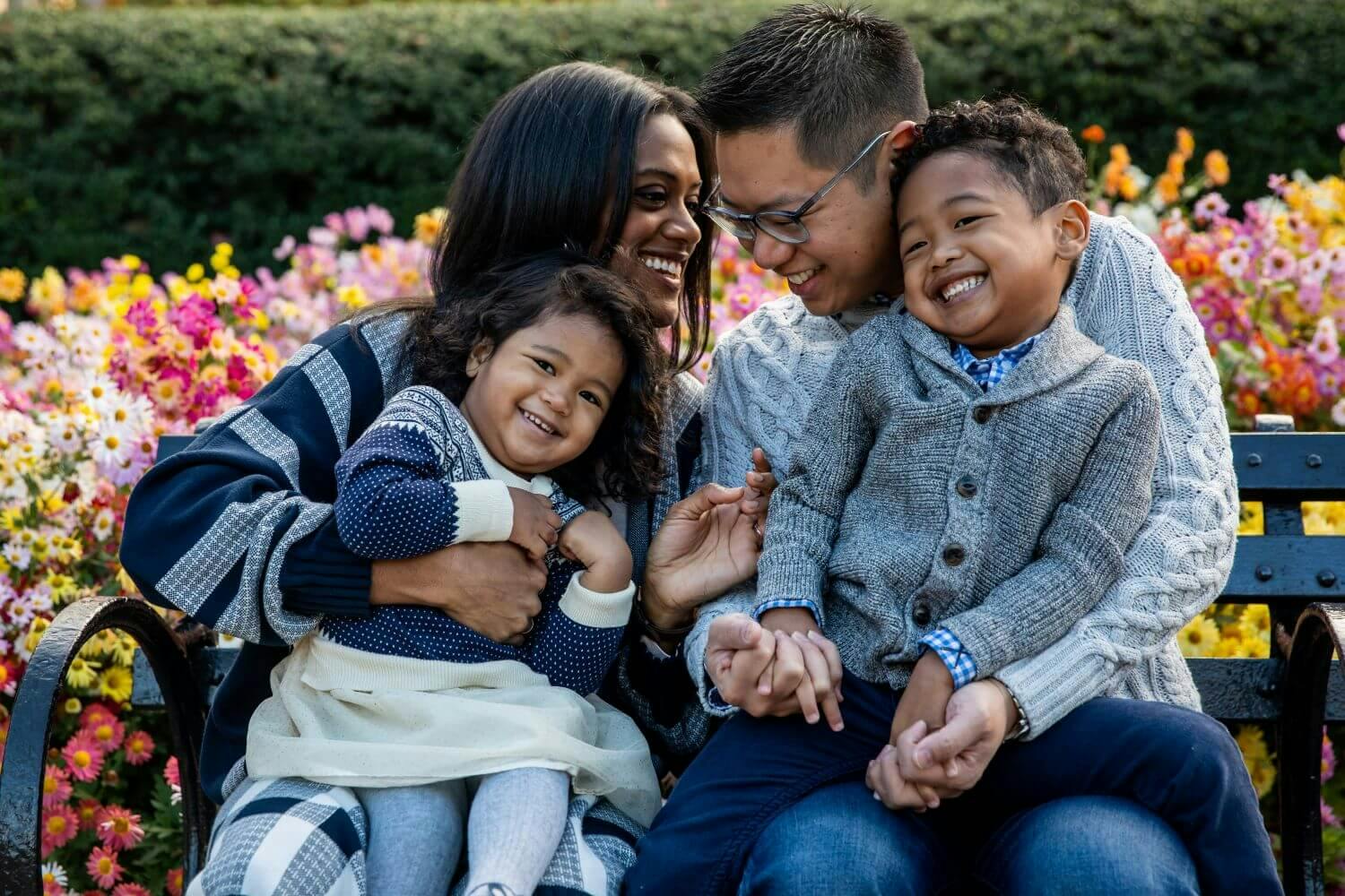 A happy family with two children smiling and hugging each other on a park bench surrounded by flowers.
