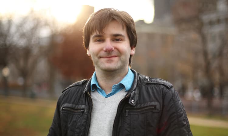 Man with a content smile wearing a jacket and blue shirt outdoors